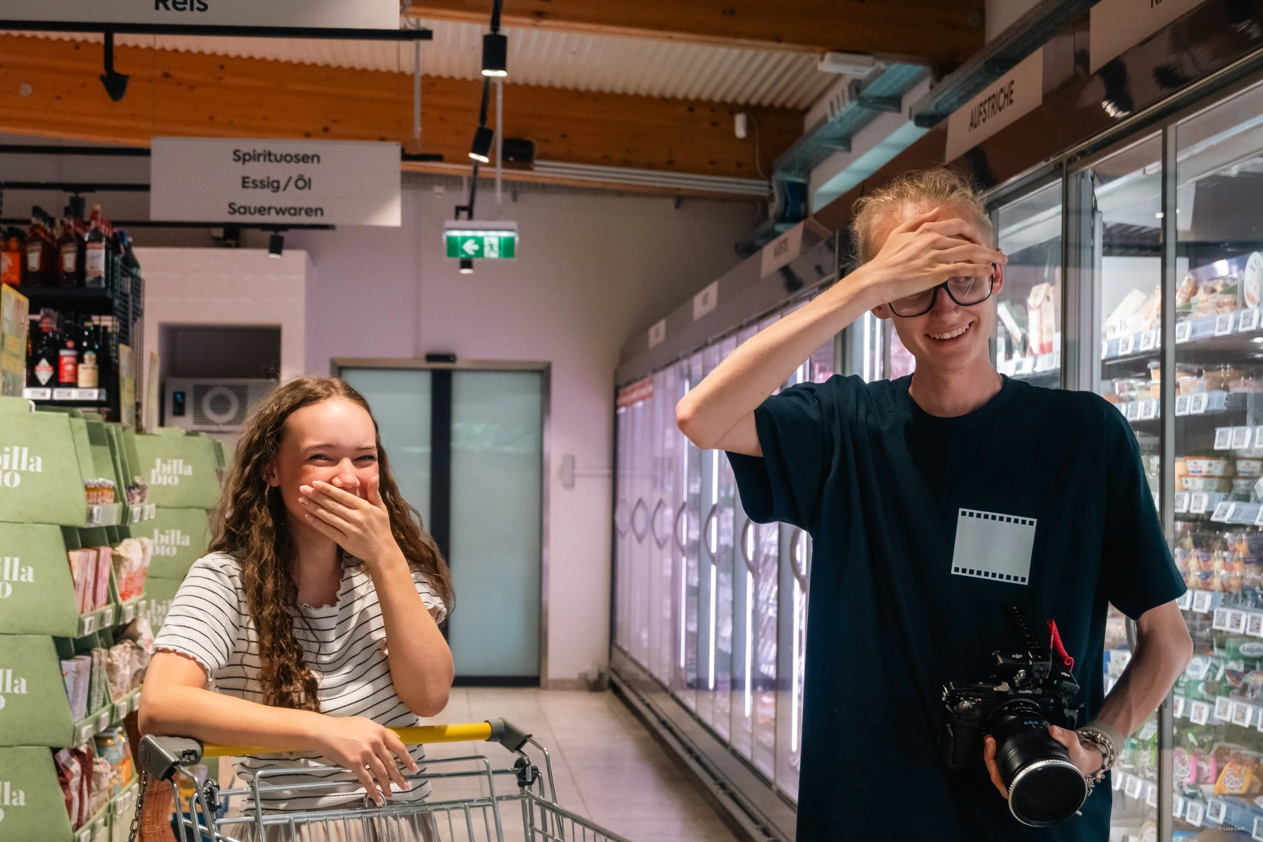 Zwei junge Menschen lachen in einem Supermarkt. Die Frau steht neben einem Einkaufswagen und hebt ihre Hand zum Gesicht, während der Mann mit Brille, schwarzem T-Shirt und Kamera eine Hand auf die Stirn legt.