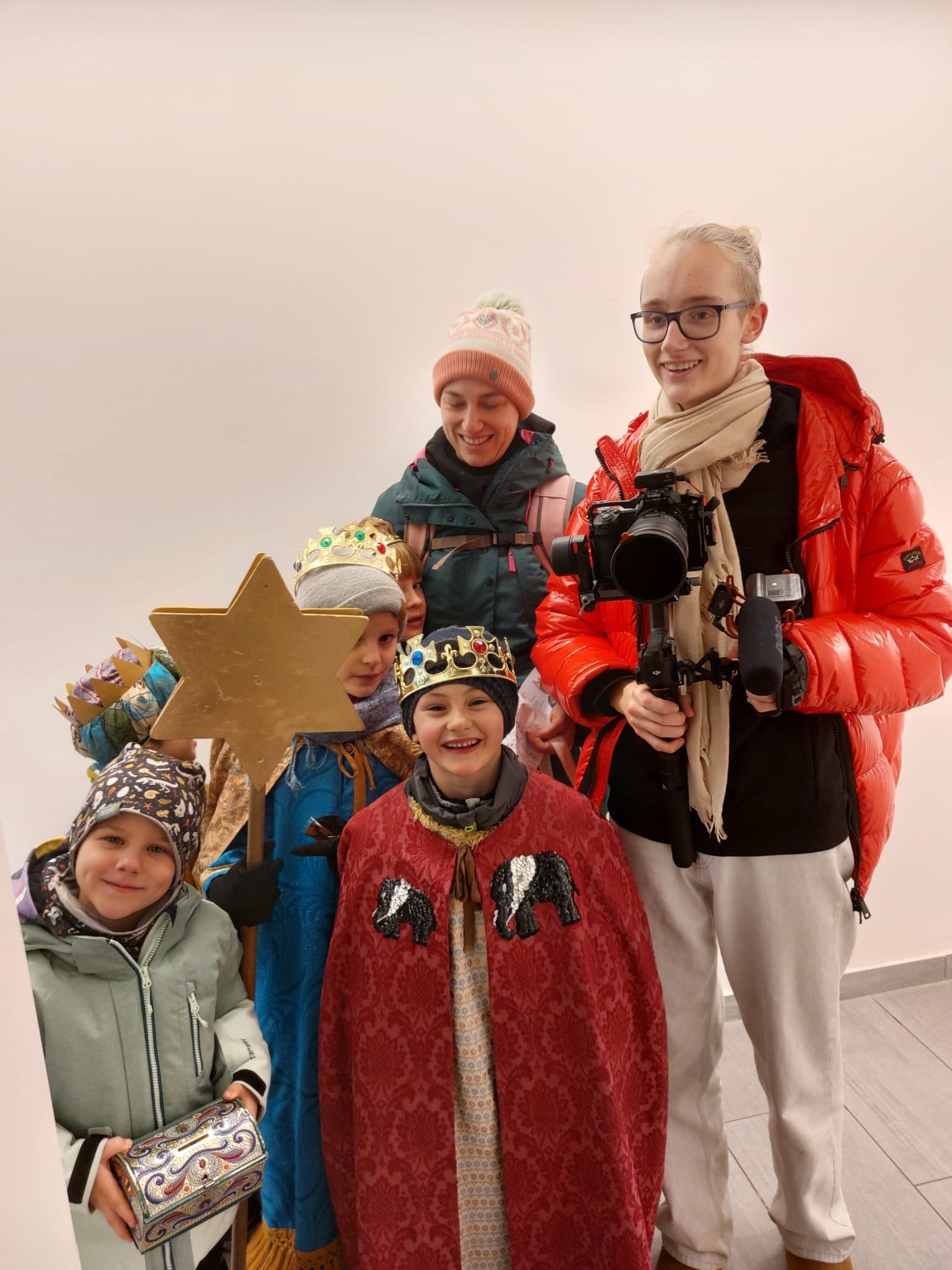 Eine Gruppe von Menschen, darunter Kinder mit Kronen, einen Erwachsenen in einem roten Mantel, macht Theater. Die Kinder tragen Kostüme, und es gibt eine goldene Stern-Requisite.