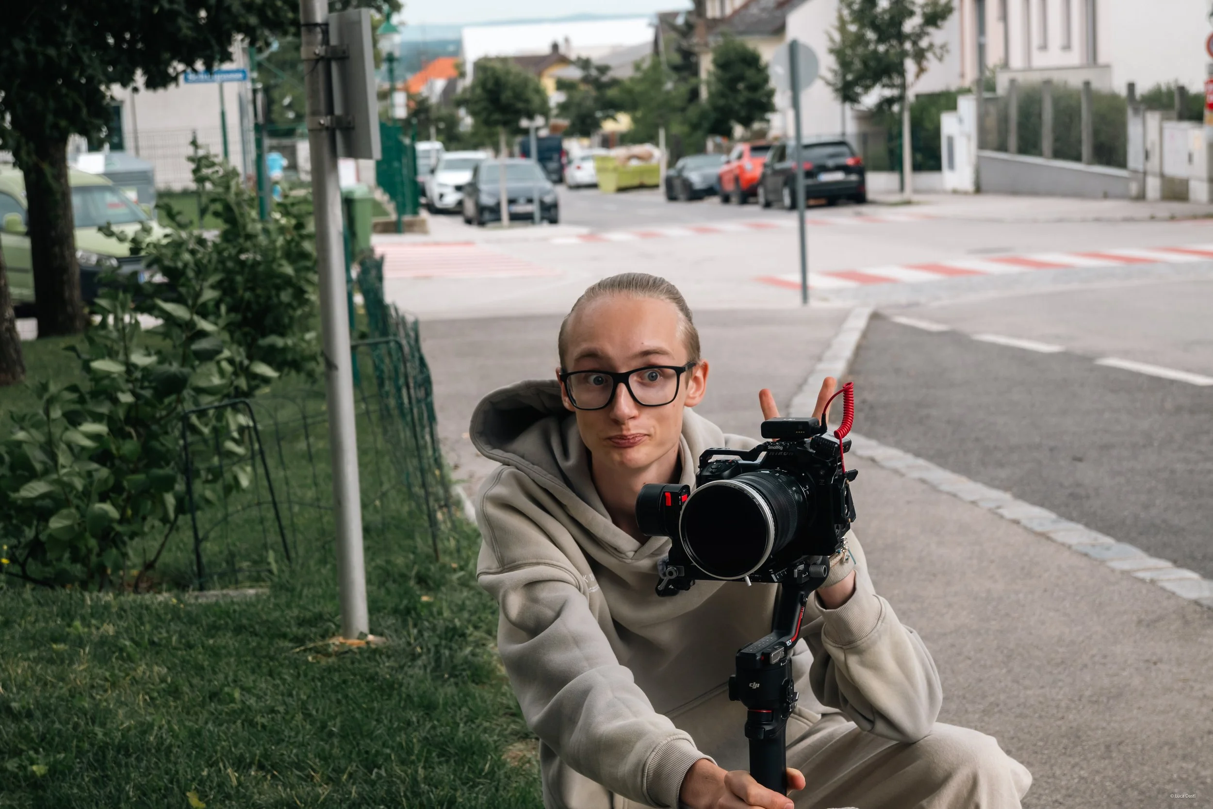 Junge Person mit Brille und Hoodie hält eine Kamera auf einem Gimbal, sitzt auf dem Bürgersteig neben Gras, im Hintergrund eine Straße und parkende Autos.