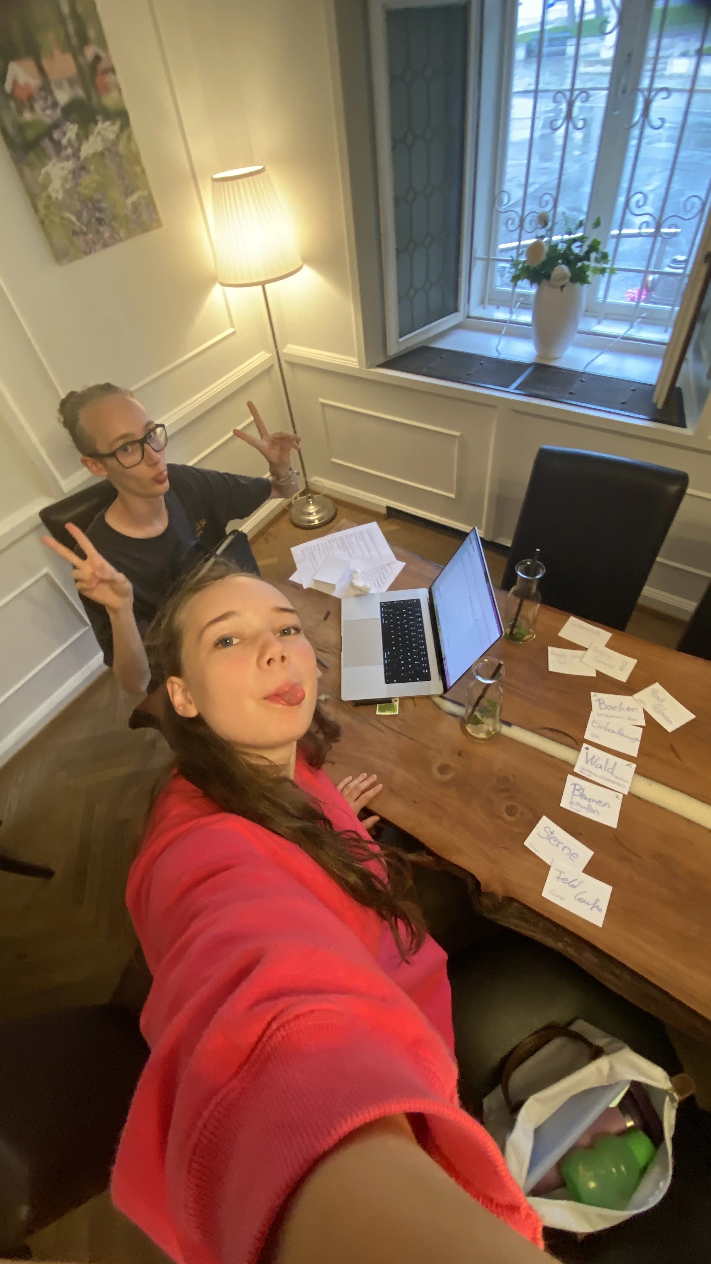 Zwei lachende junge Frauen nehmen ein Selfie in einem hell erleuchteten Raum auf, der eine große Holztisch mit Papieren, Laptops und Wasserflaschen hat, vor einem Fenster mit Gitter und einer weißen Vase mit Blumen.