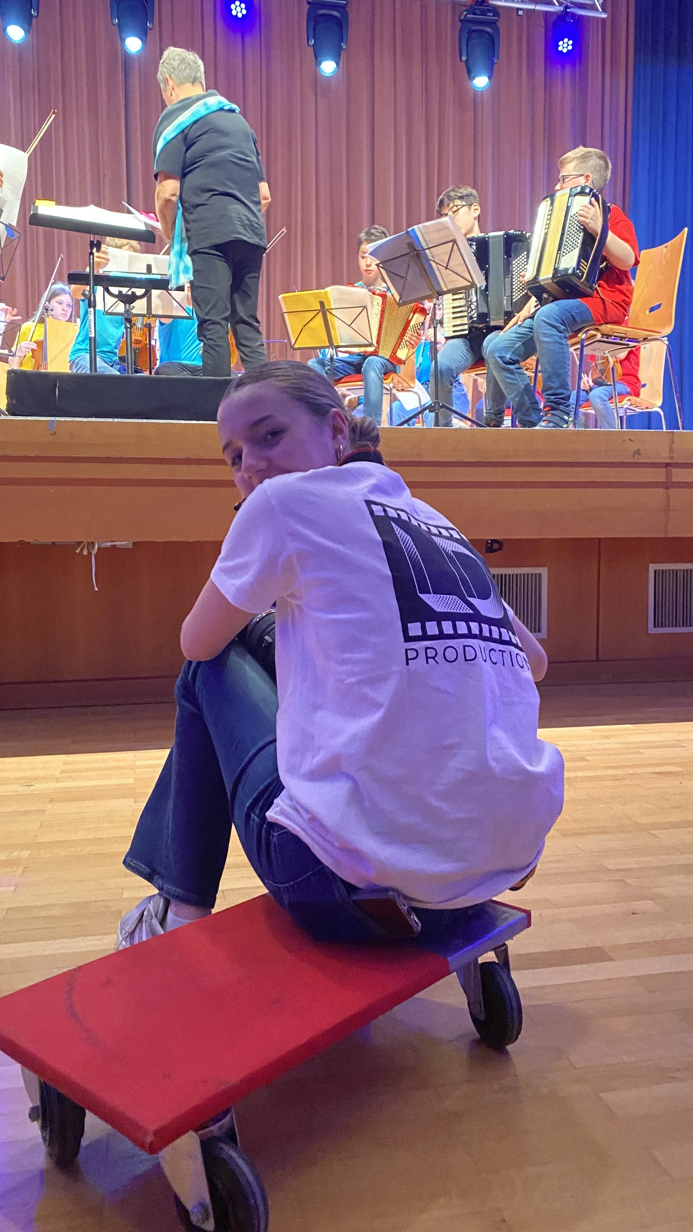 Jugendliche sitzt auf einem roten Rollbrett vor einer Bühne mit einem Orchester, das auf Musikstücke vorbereitet ist.
