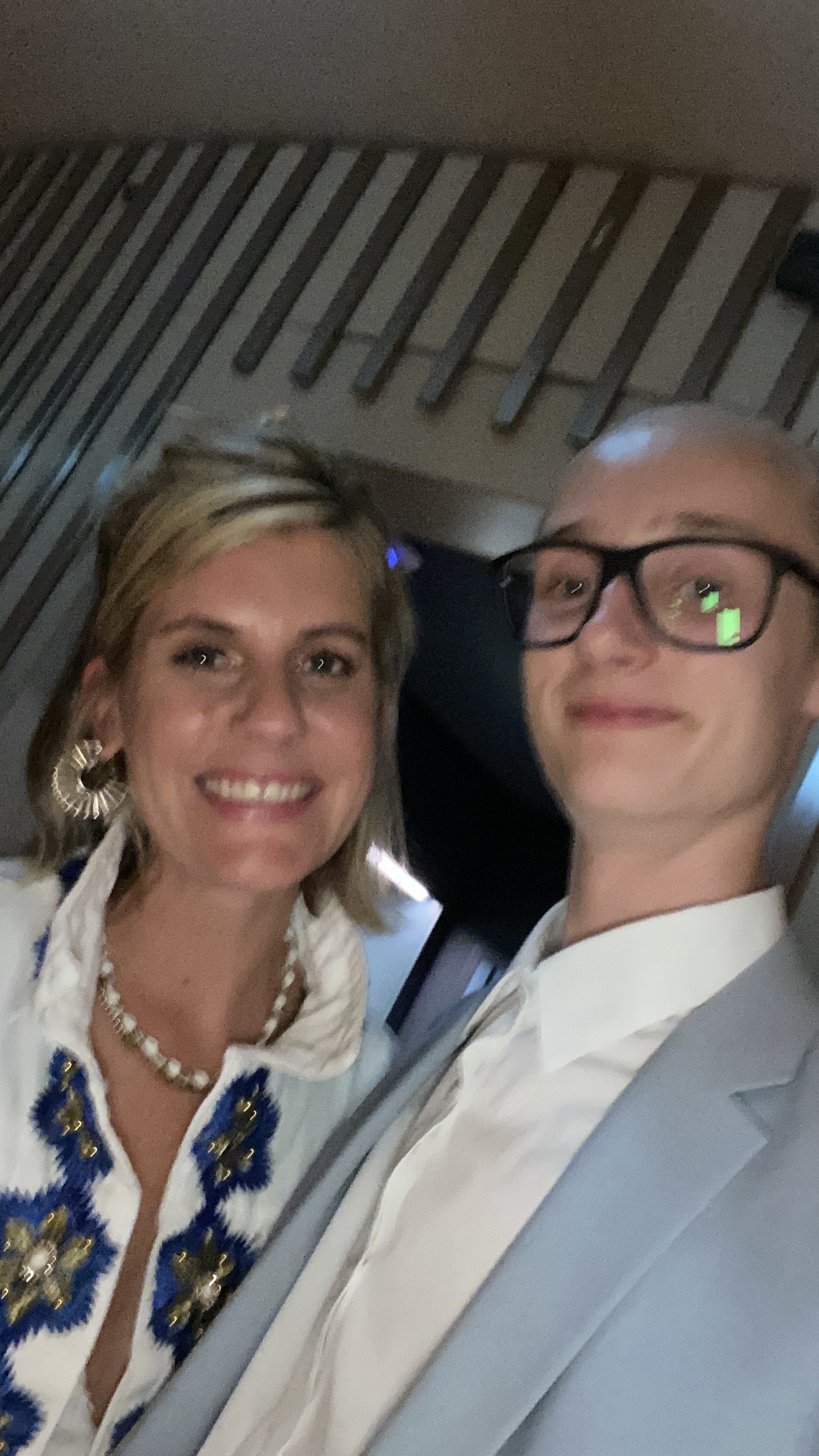 Ein Mann und eine Frau posieren für ein Selfie, beide lächeln. Der Mann trägt eine Brille und ein helles Jackett, die Frau trägt Ohrringe, eine Halskette und ein Oberteil mit blauen und goldenen Verzierungen.