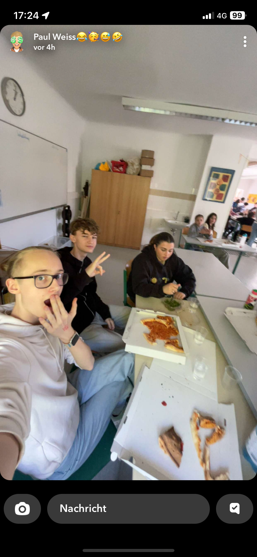 Jugendliche sitzen an einem Tisch in einem Klassenzimmer, essen Pizza, trinken Wasser und fotografieren sich selbst, während auf dem Tisch ein halb gegessenes Pizzastück liegt.