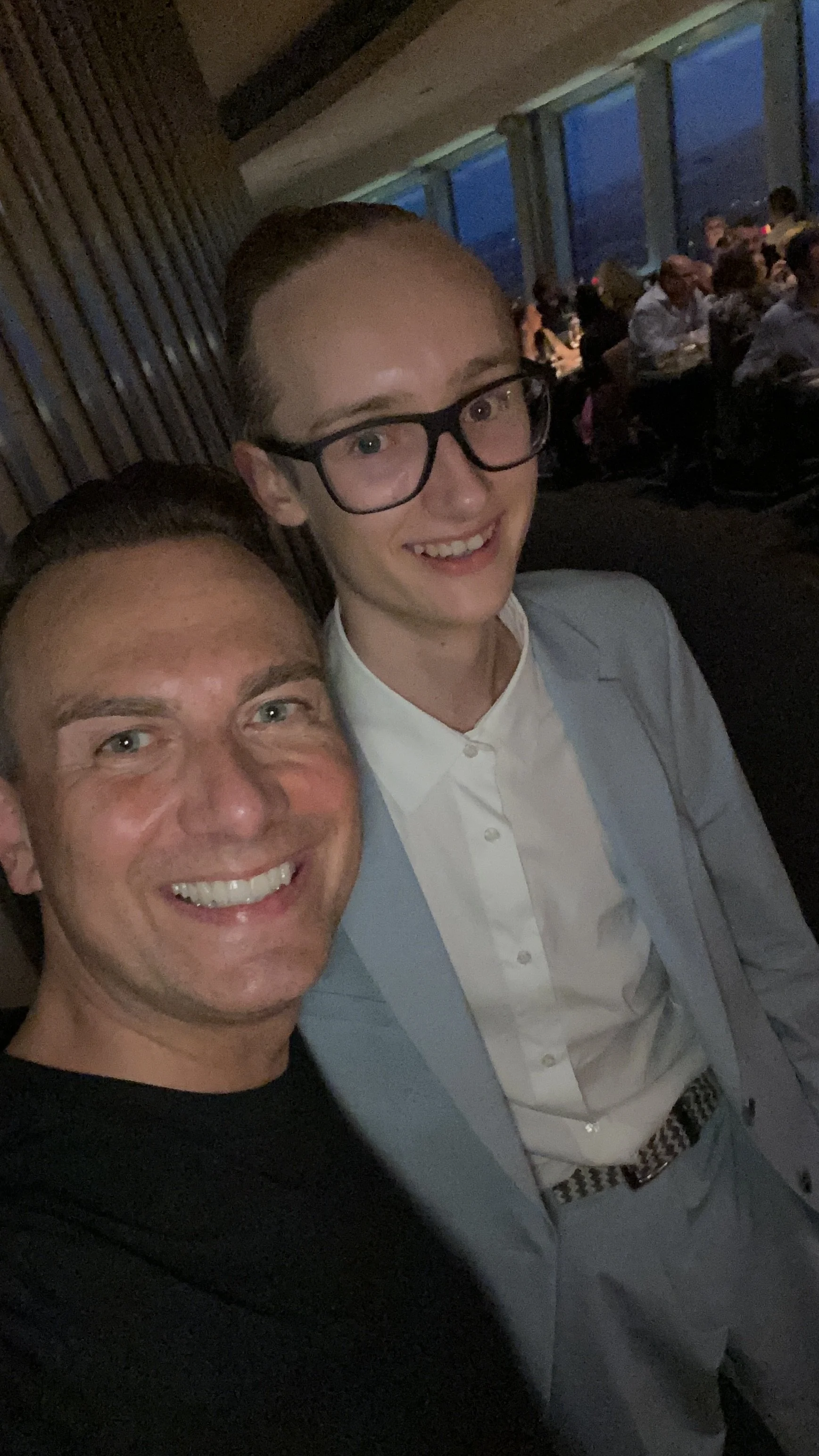 Zwei Männer, die auf einer Party oder in einem Restaurant gelacht und fotografiert werden. Der Mann im Vordergrund trägt ein schwarzes Hemd, der Mann im Hintergrund trägt eine helle Anzugjacke, weißes Hemd und hat Brille.