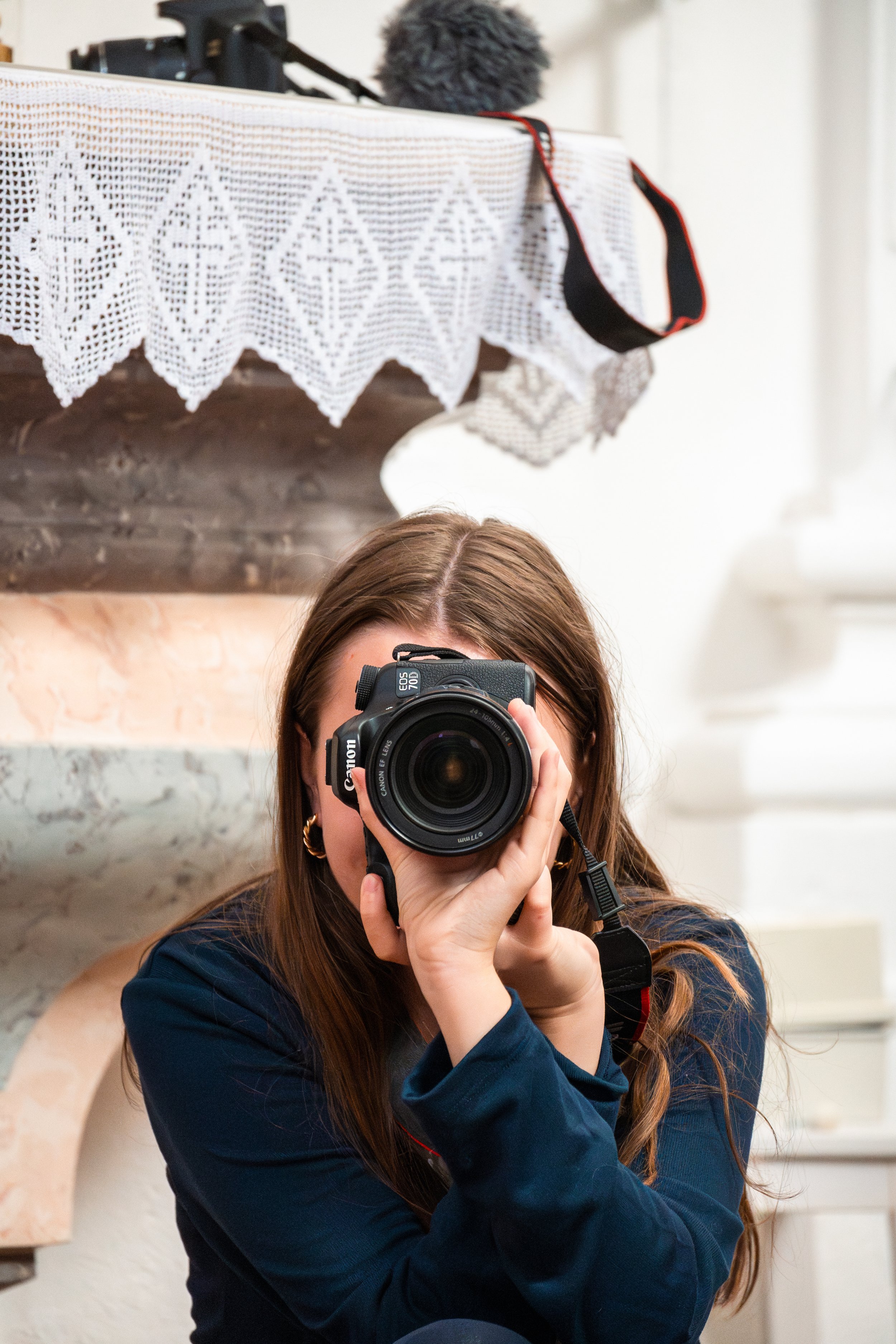 Eine Frau mit braunen Haaren, die eine Kamera in der Hand hält und in die Linse schaut, während sie fotografiert. Im Hintergrund ist ein Kamin und eine Tischdecke mit Spitze.