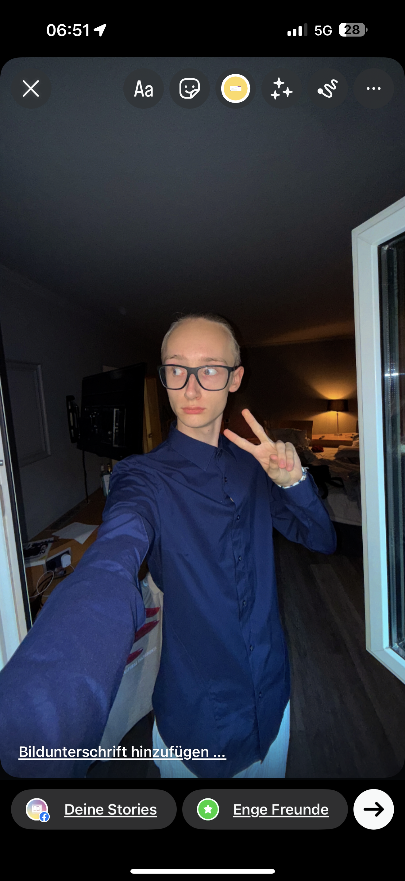 Ein junger Mann mit Brille trägt ein dunkelblaues Hemd und macht eine Peace-Geste mit der rechten Hand, während er eine Selfie-Fotografie in einem dunklen Raum aufnimmt.