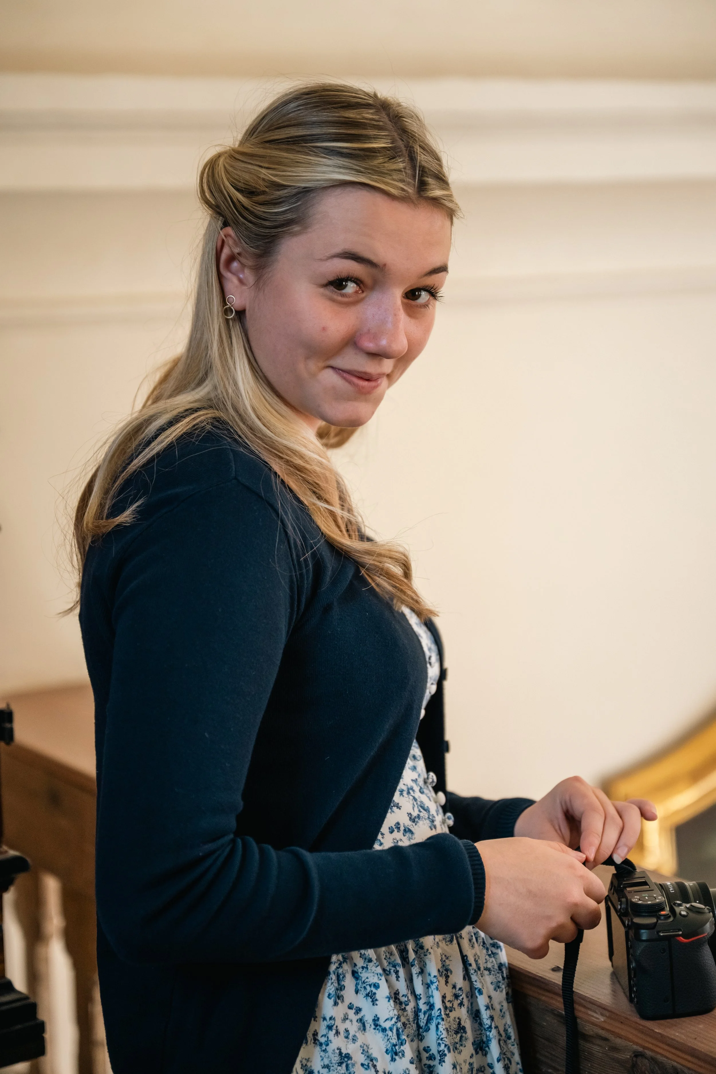 Jugendliche Frau steht an einem Tisch, hält eine Kamera in den Händen und lächelt leicht.