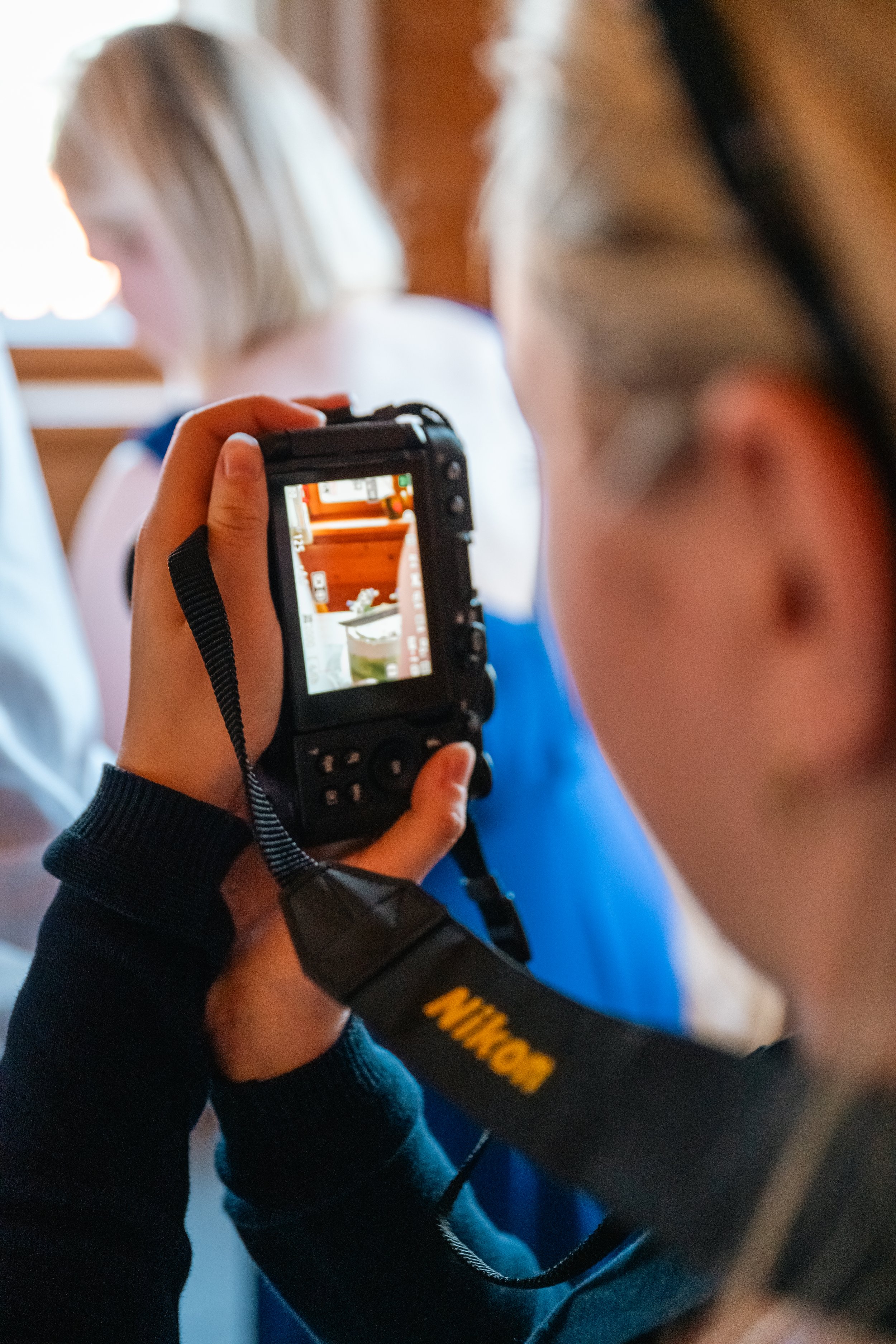 Eine Person fotografiert mit einer digitalen Nikon-Kamera eine Szene, die auf einem Bildschirm der Kamera sichtbar ist. Im Hintergrund ist eine andere Person mit blondem Haar, leicht unscharf, zu sehen.
