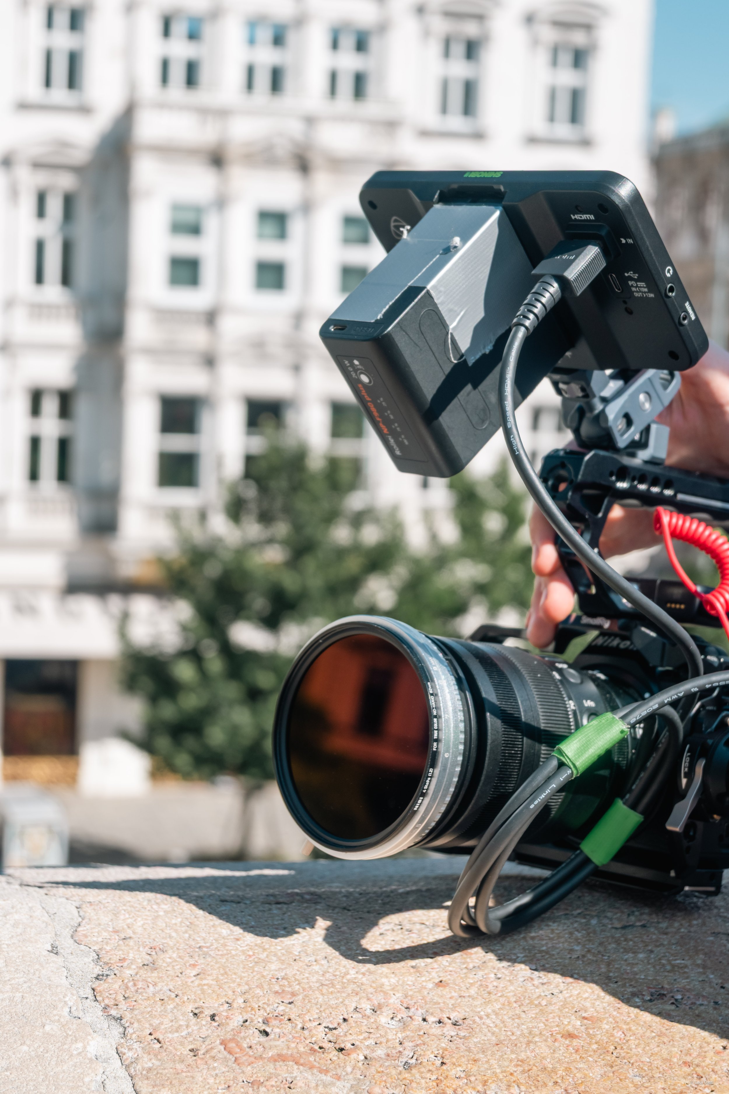 Kamera mit externem Monitor und Akku auf einer Terrasse im Freien, im Hintergrund ein mehrstöckiges Gebäude mit weißen Fassaden.