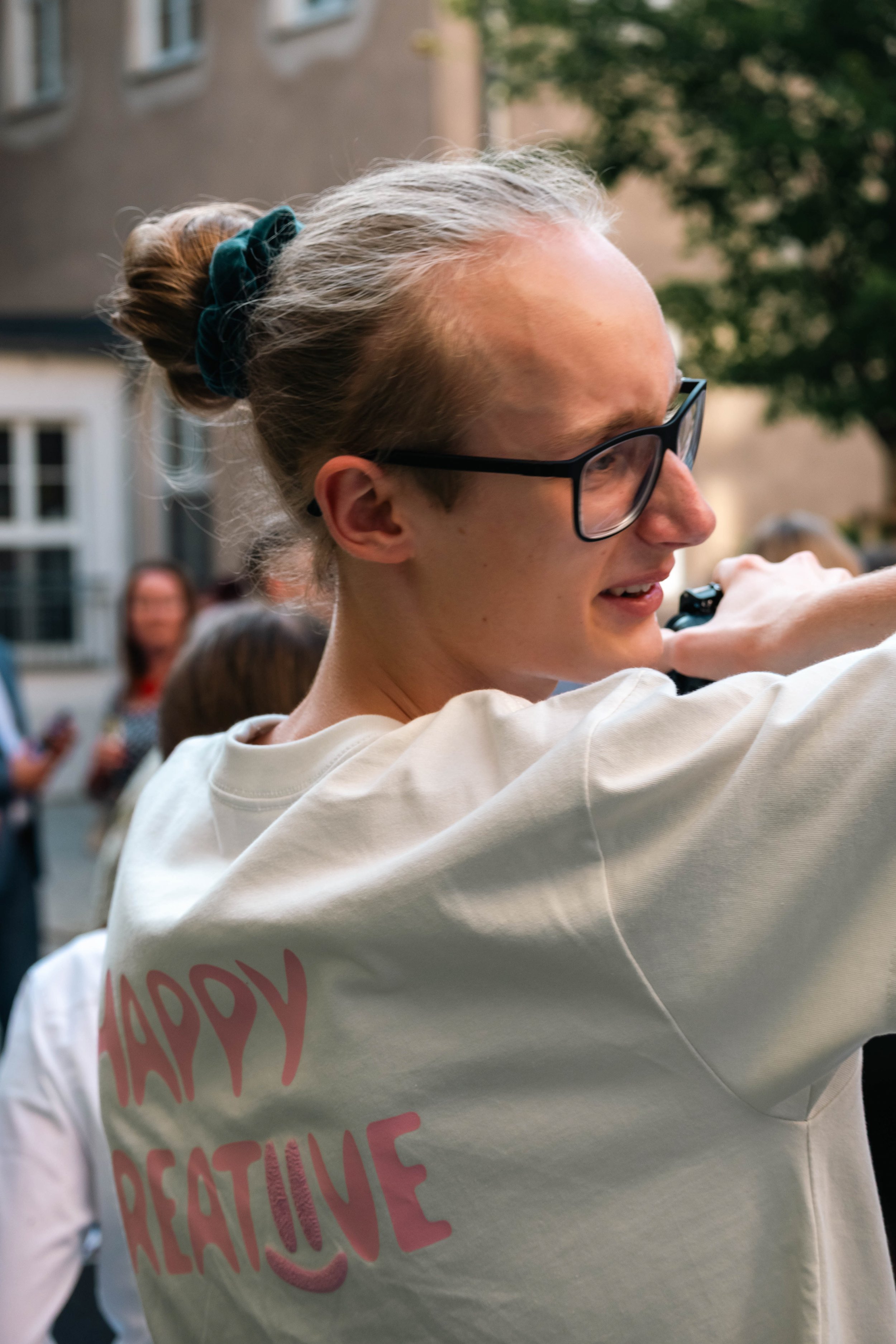 Junge Frau mit Brille und Dutt, die ein T-Shirt trägt, auf dem steht 'HAPPY CREATIVE', nimmt ein Foto oder Video mit einer Kamera in einer Gruppe im Freien auf einer Straße auf, im Hintergrund sind andere Personen und ein Gebäude.