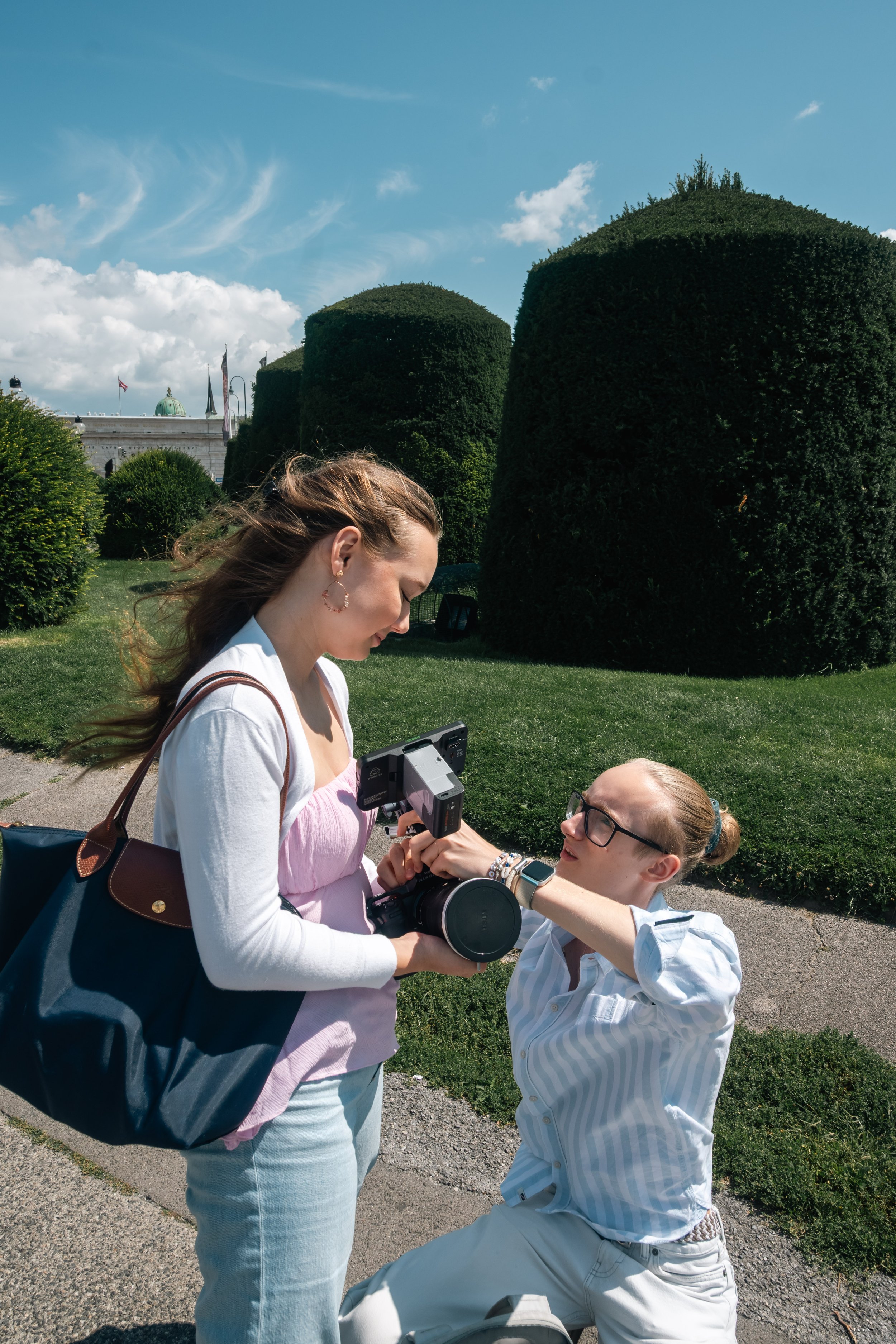 Zwei Frauen auf einer Wiese, eine sitzt mit einem Verlobungsring an ihrem Finger und die andere steht vor ihr, zeigt auf den Ring und hält eine Kamera. Im Hintergrund sind große, grüne, kugelförmige Büsche und ein blauer Himmel mit Wolken.