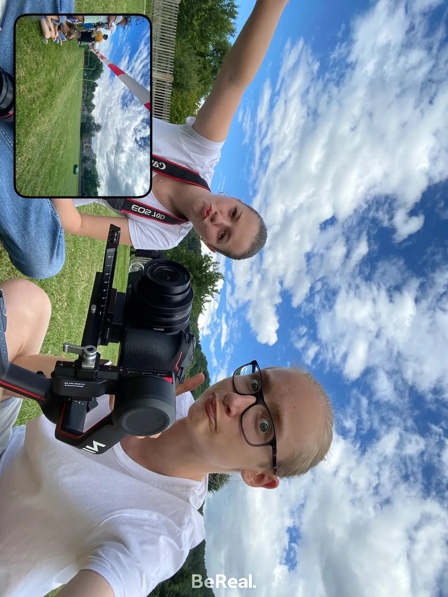Zwei junge Menschen, ein Mann mit Brille und ein Frau, machen ein Selfie im Freien unter einem blauen Himmel mit Wolken. Im Hintergrund sind Bäume und eine Grünfläche sichtbar.