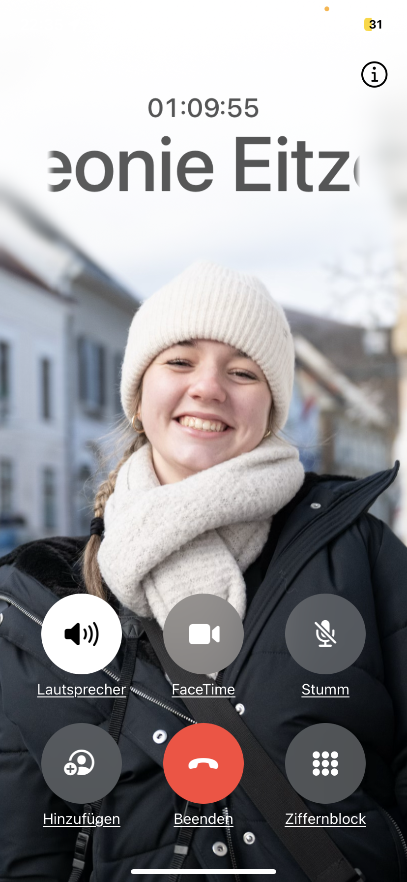 Eine lachende junge Frau beim Videoanruf im Freien bei kaltem Wetter, trägt einen weißen Wollmütze und Schal, schwarzer Winterjacke, im Hintergrund sind Häuser und Bäume sichtbar.