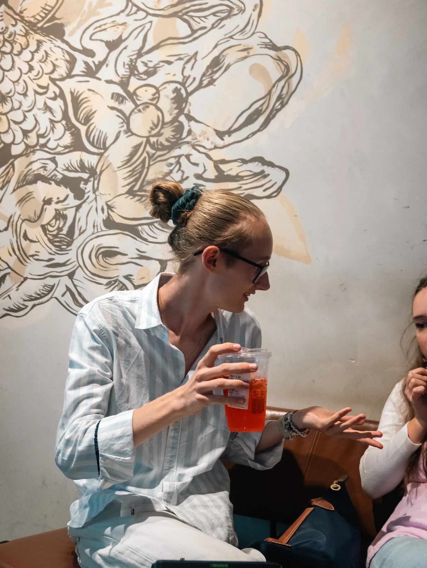 Eine junge Frau mit Brille, in einem weißen gestreiften Hemd, sitzt auf einer Bank und hält ein Getränk in der Hand, während sie mit einer anderen Person spricht. Im Hintergrund eine Wand mit Wandkunst. Die Szene scheint in einem Café oder Restaurant