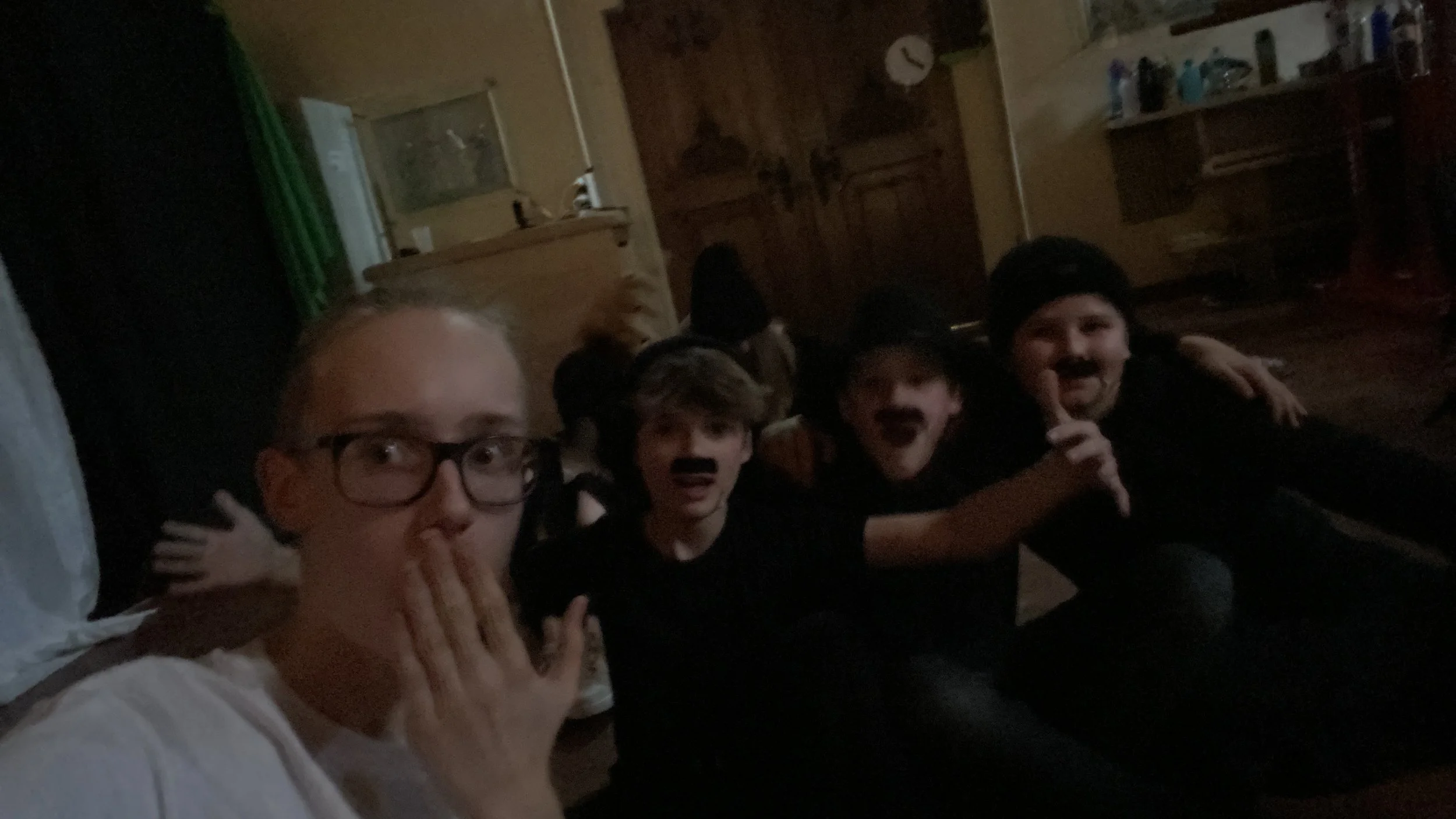 Vier Jungen sitzen auf dem Boden in einem Zimmer und spielen, alle haben schwarze Schnurrbärte aufgemalt.