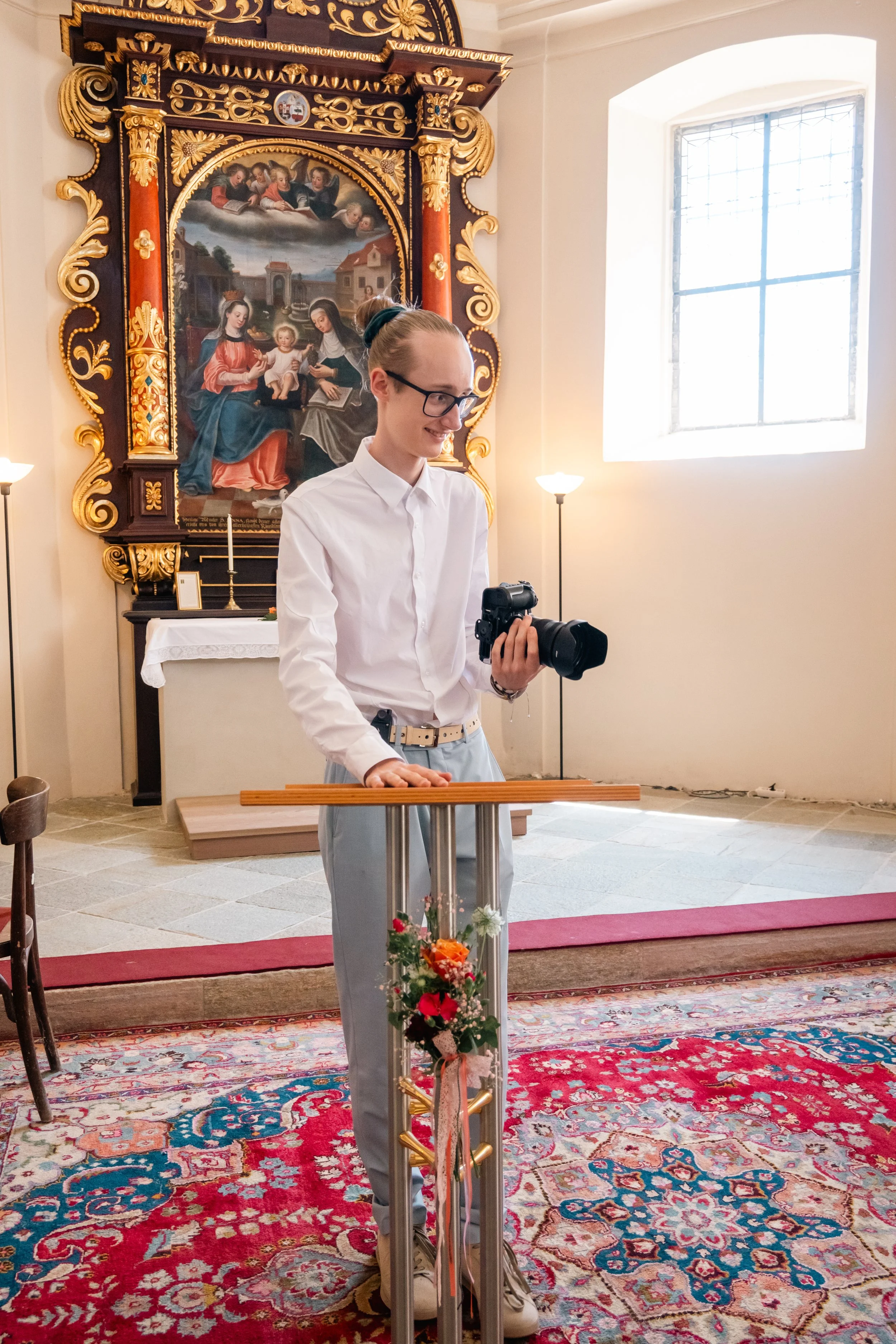 Eine junge Frau steht an einem Mikrofon in einer Kirche, hält eine Kamera in der Hand und lächelt. Im Hintergrund ist ein barockes Altarbild mit religiöser Szene, ein Fenster und Stehlampen.