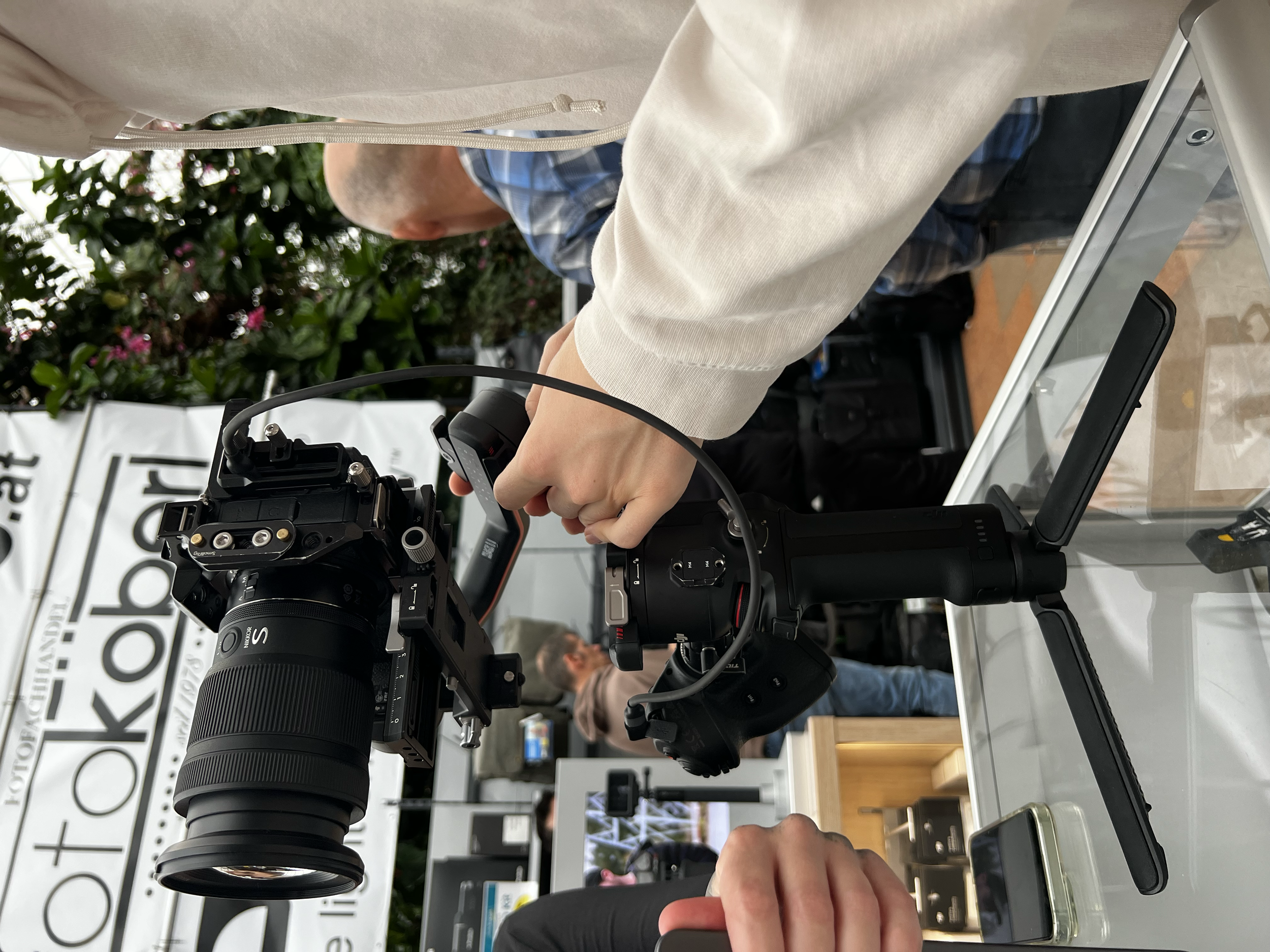 Nahaufnahme einer Kamera, die auf einem Metallständer montiert ist, während eine Person die Kamera bedient, im Hintergrund Pflanzen und eine Messeumgebung mit anderen Personen und einem Banner.