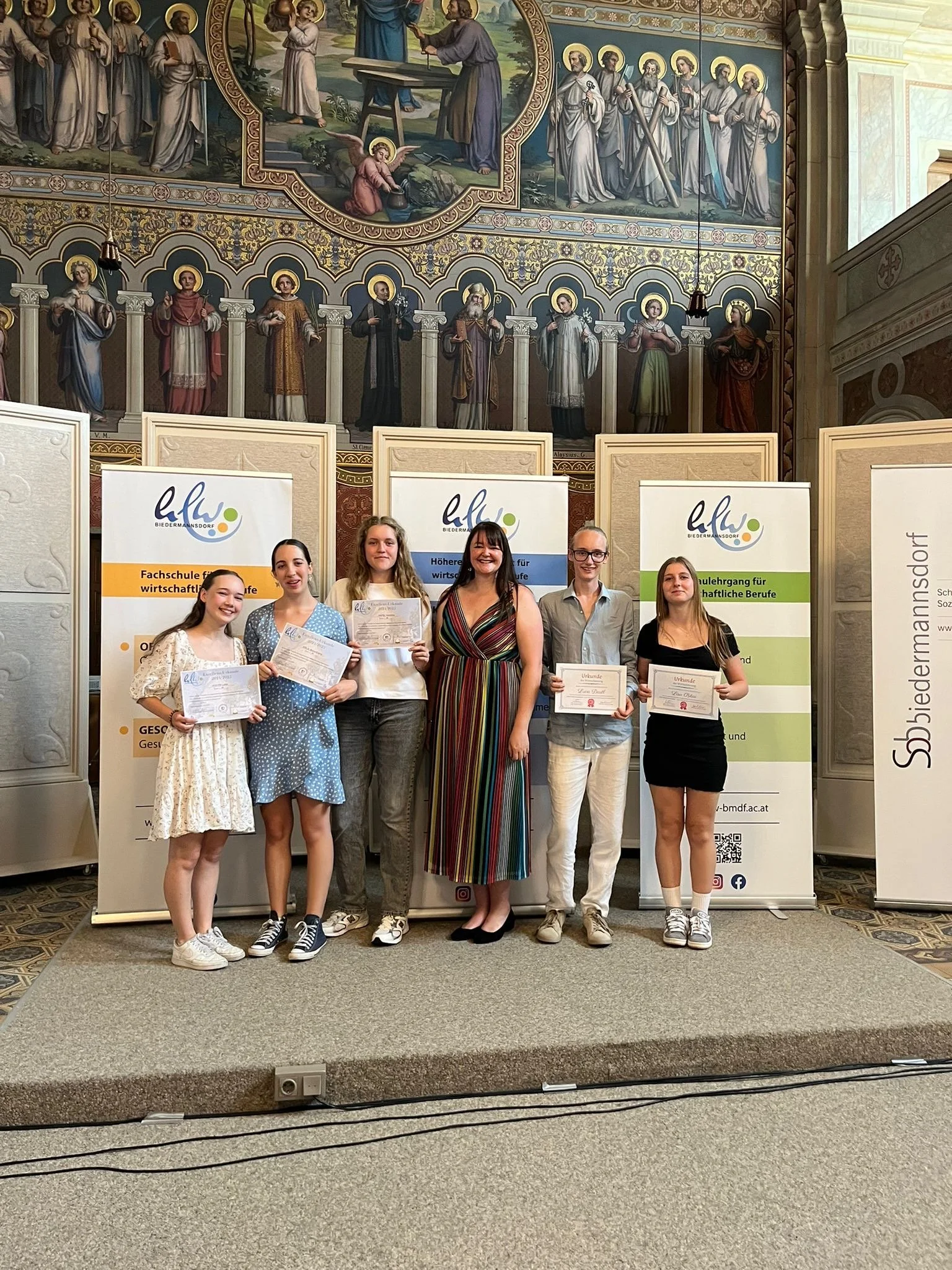 Eine Gruppe junger Frauen und eine ältere Frau stehen auf einer Bühne und halten Urkunden in einem Raum mit religiösen Wandmalereien im Hintergrund. Es handelt sich wahrscheinlich um eine Preisverleihung oder Zeremonie.