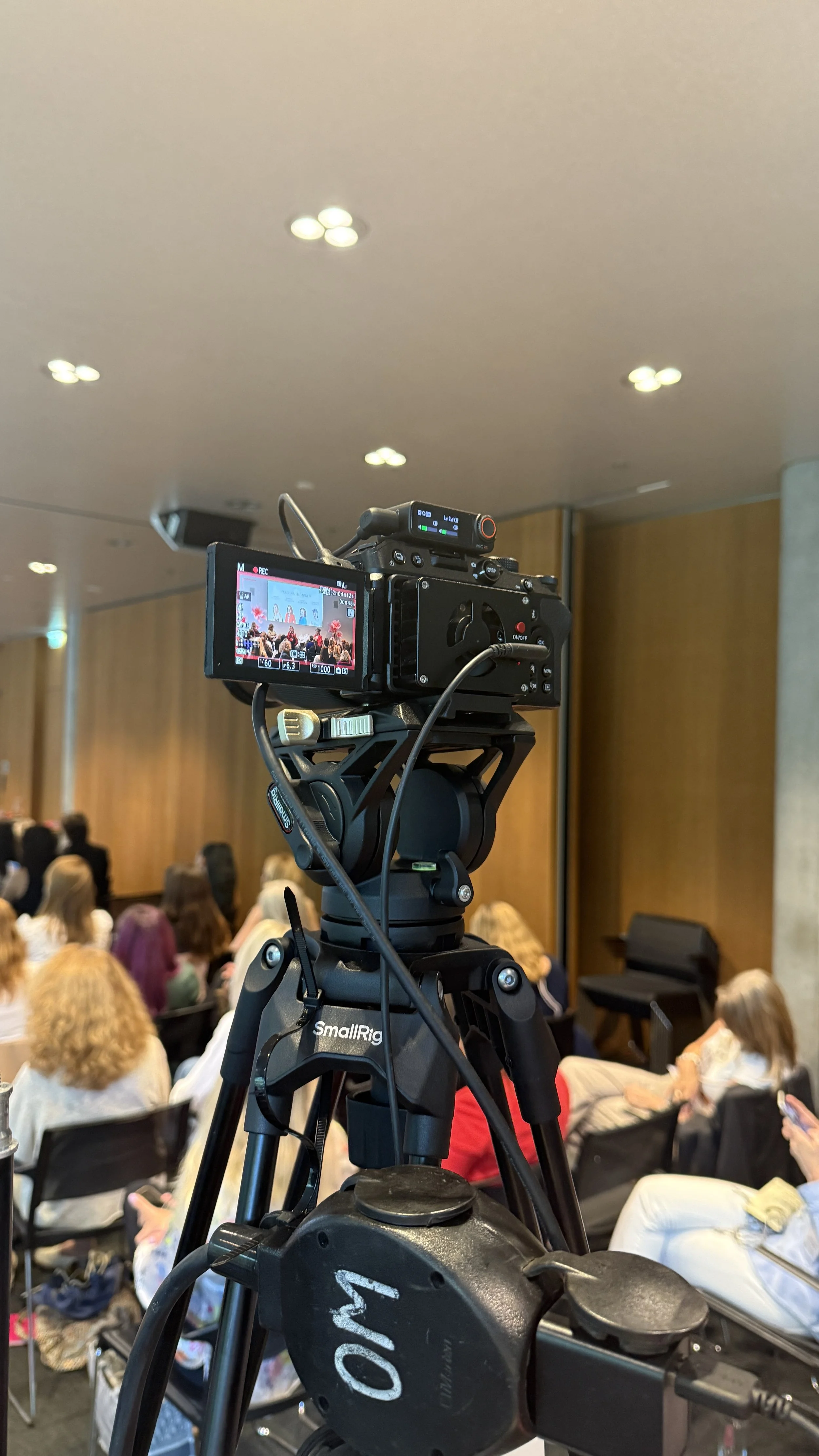 Eine Kamera auf einem Stativ filmt eine Konferenz oder Veranstaltung in einem Raum mit Publikum.