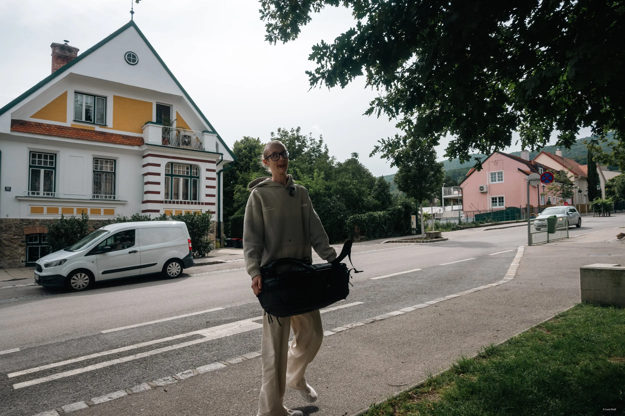 Eine Person in Freizeitkleidung trägt einen Rucksack mit einem Hund und geht auf einem Gehweg, im Hintergrund sind bunte Häuser und ein parkender Van zu sehen, mit Bäumen über dem Weg.