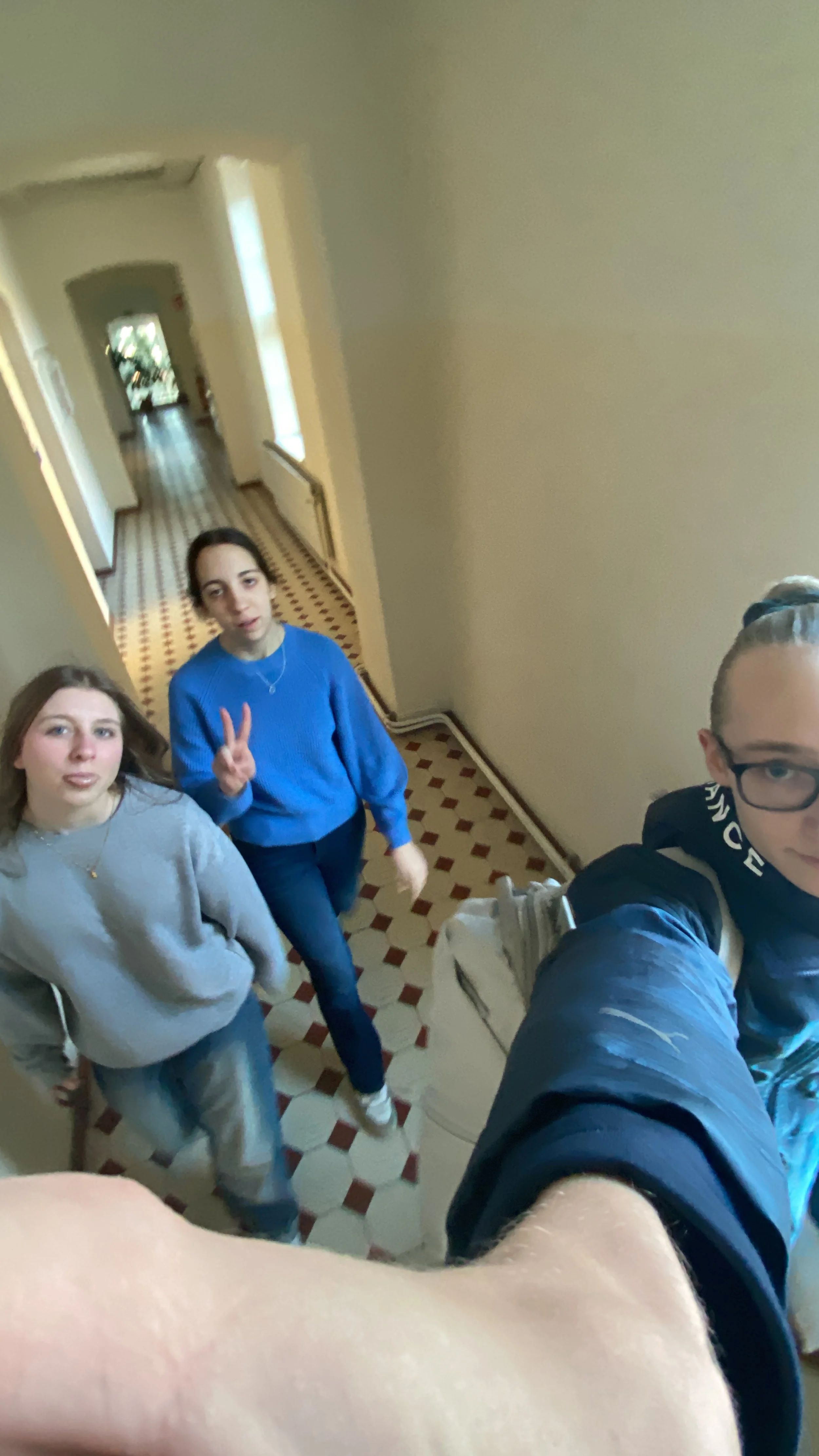 Drei junge Frauen stehen in einem Flur und fotografieren die Kamera, eine macht ein Peace-Zeichen, im Vordergrund ist der Arm eines Fotografen zu sehen.