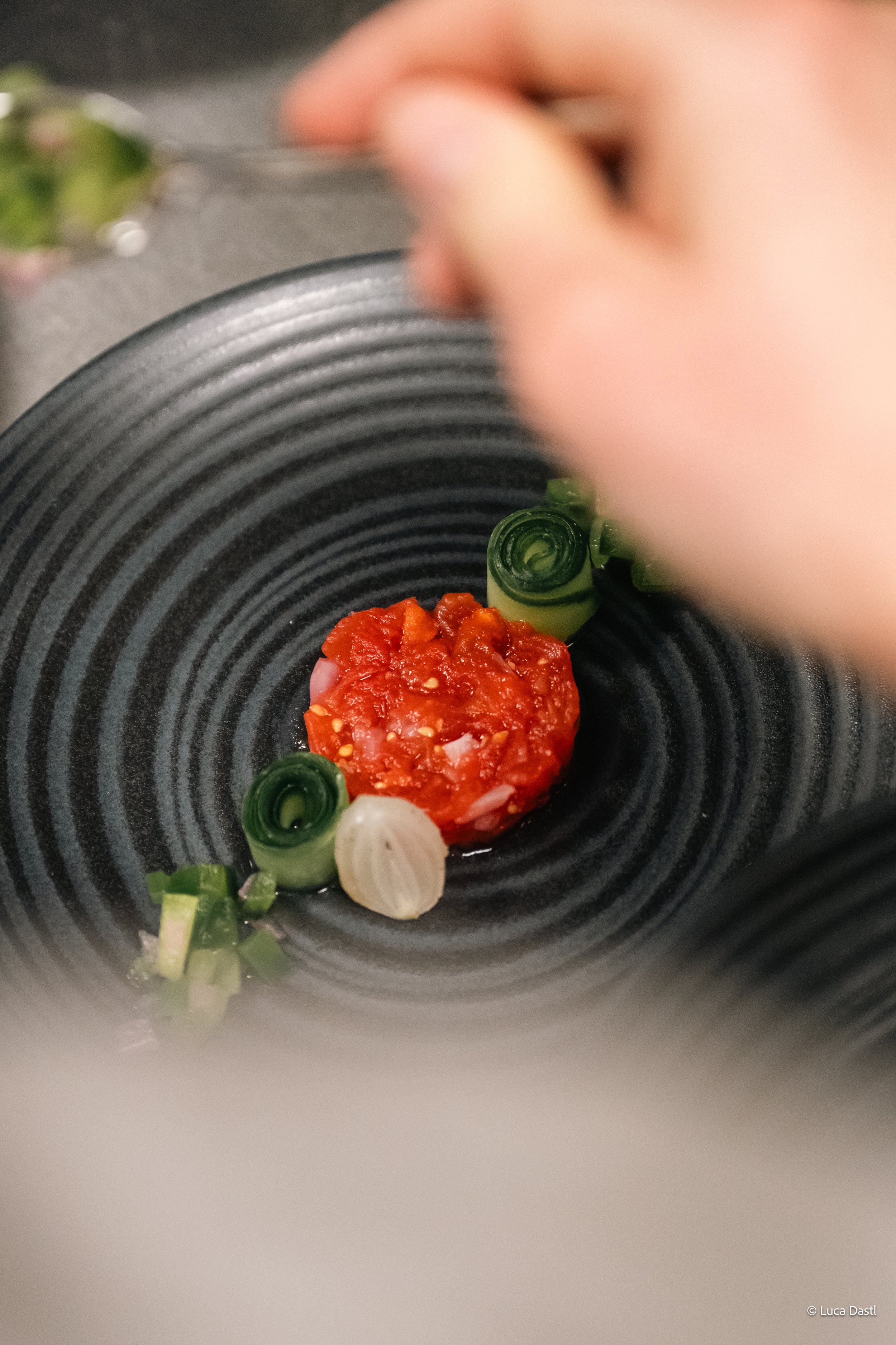 Nahaufnahme eines schwarzen Tellern mit gewürzter roter Tomatenmischung, grünen und weißen Frühlingszwiebeln, beim Garnieren. Im Hintergrund unscharf eine Hand, die mit einer Gabel eine Tomate hält.