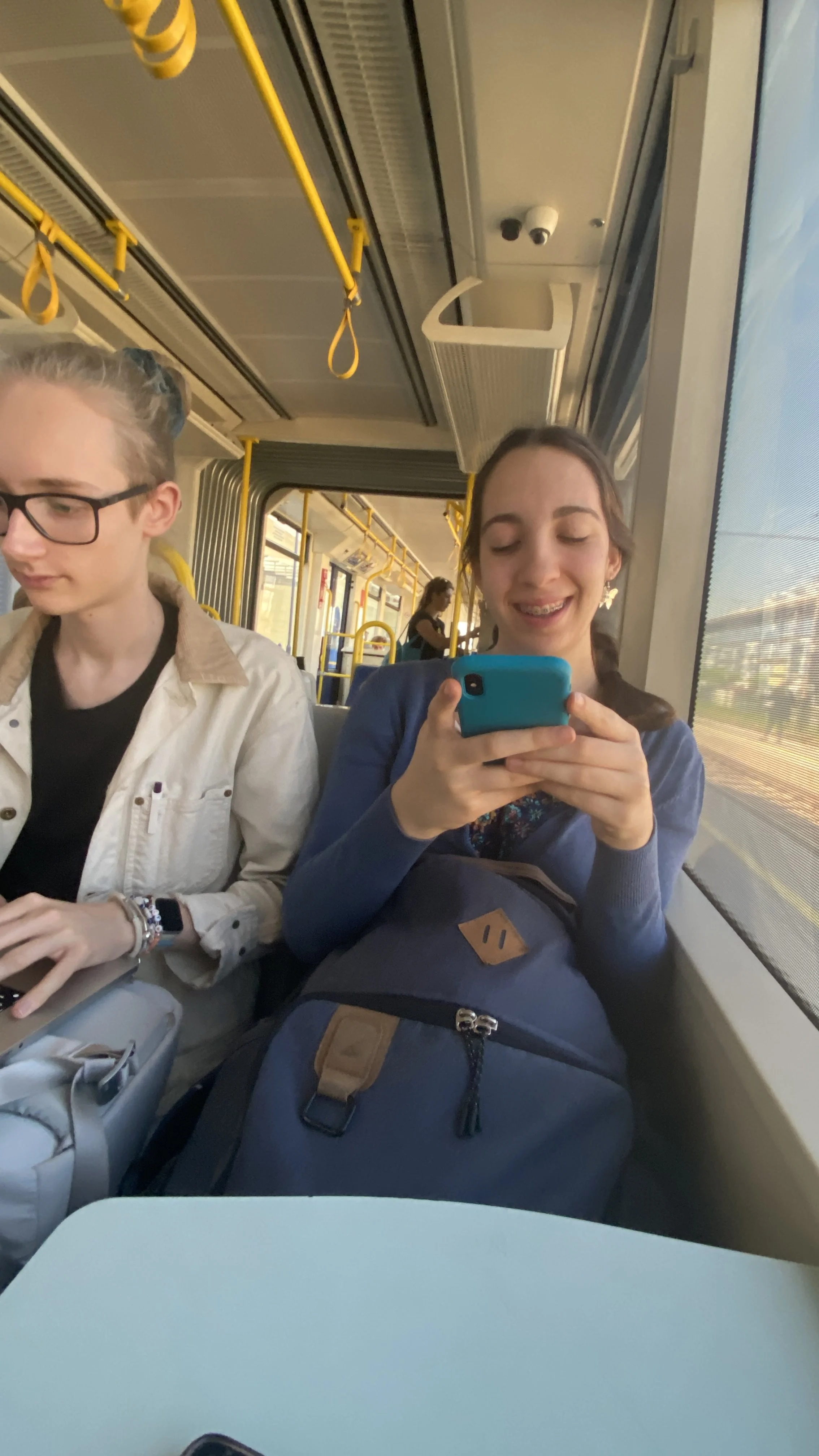 Zwei junge Frauen sitzen in einem öffentlichen Verkehrsmittel. Eine liest auf ihrem Smartphone, die andere schaut lächelnd auf ihr Telefon. Im Hintergrund sind weitere Fahrgäste und das Innere des Busses sichtbar.