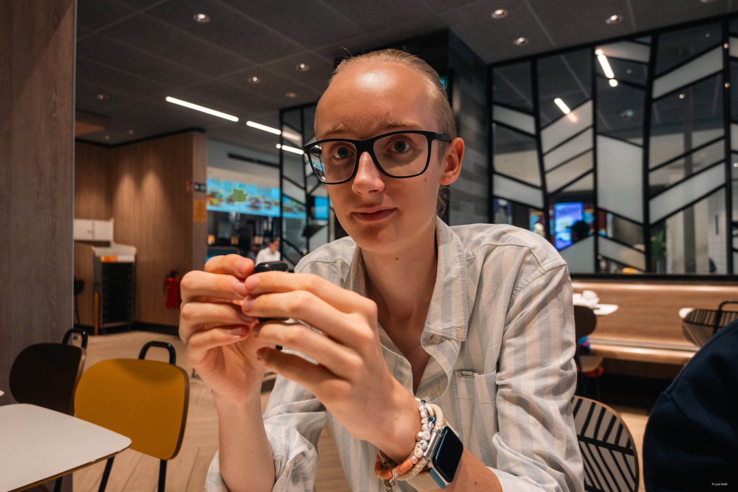 Junge Frau mit Brille und Smartwatch sitzt in einem Restaurant und hält ihre Hände zusammen, Blick nach vorne.