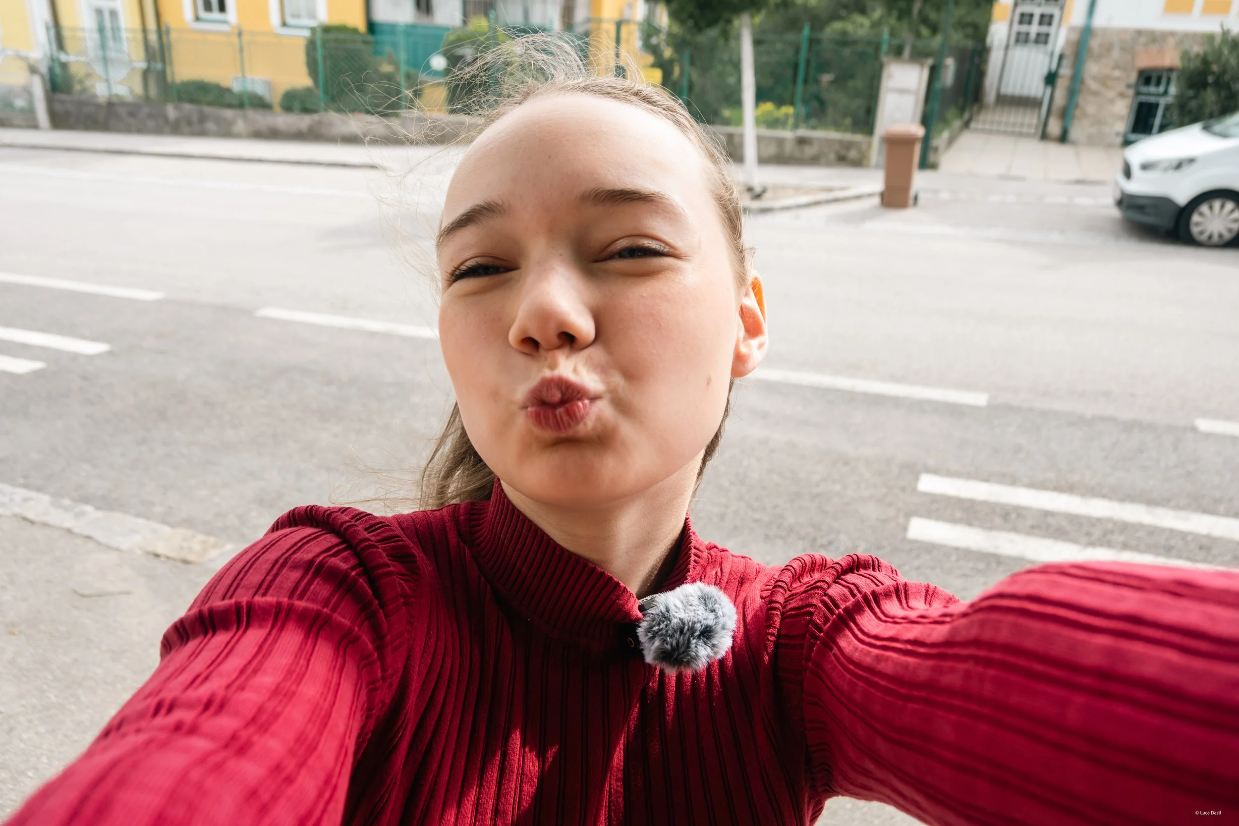 Junge Frau macht einen Selfie-Foto auf der Straße, trägt eine rote Jacke, macht Kussmund, und hat eine graue Plüschpinsel am Kragen.