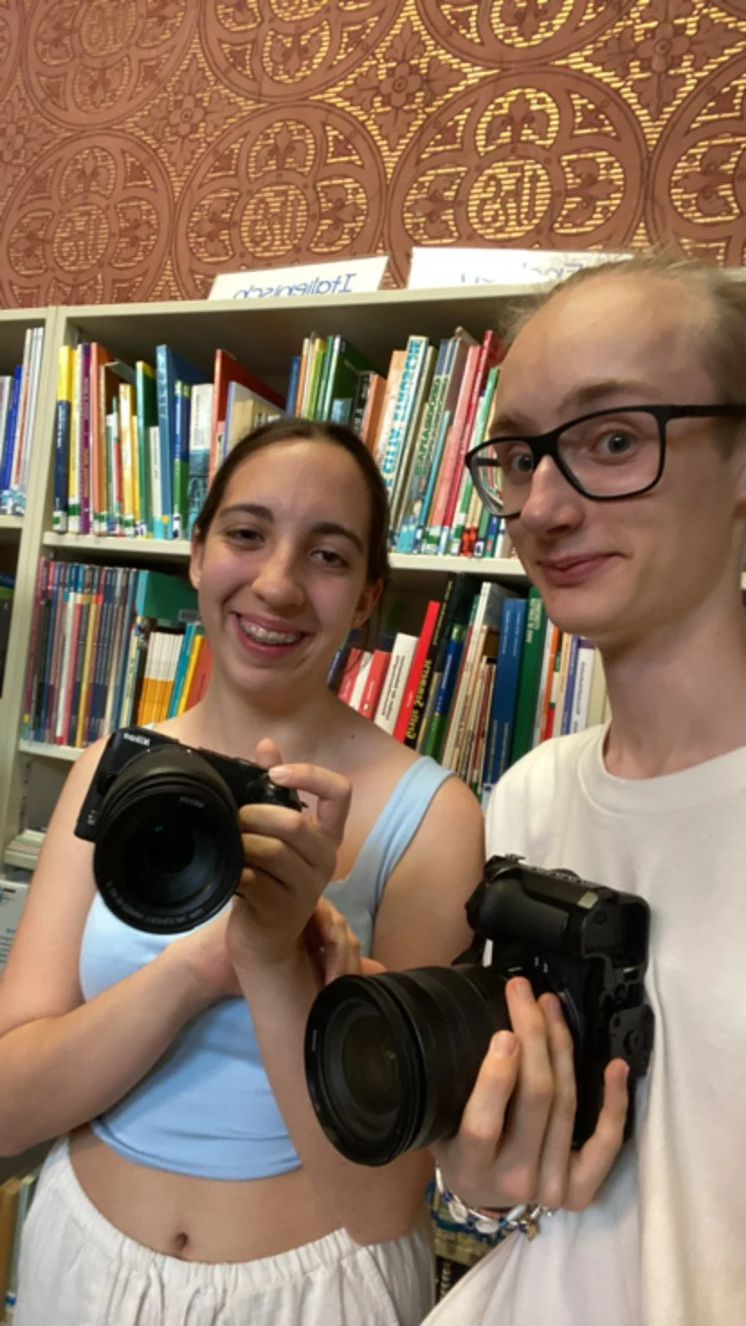 Zwei Jugendliche in einer Bibliothek, die beide Kameras in den Händen halten und für einen Selfie-Foto posieren.