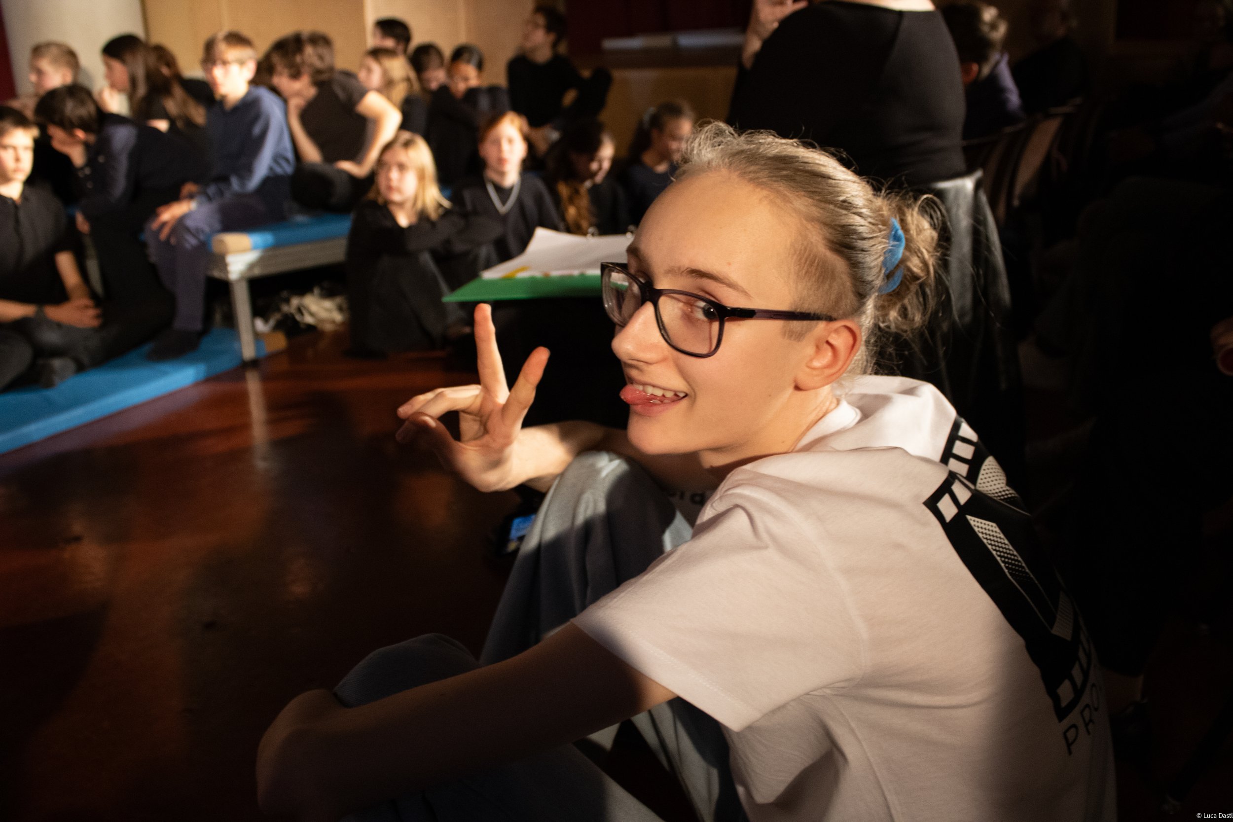 Ein lachender junger Mensch mit Brille macht ein Peace-Zeichen und stößt im Hintergrund auf eine Gruppe von Menschen in einem dunklen Raum.
