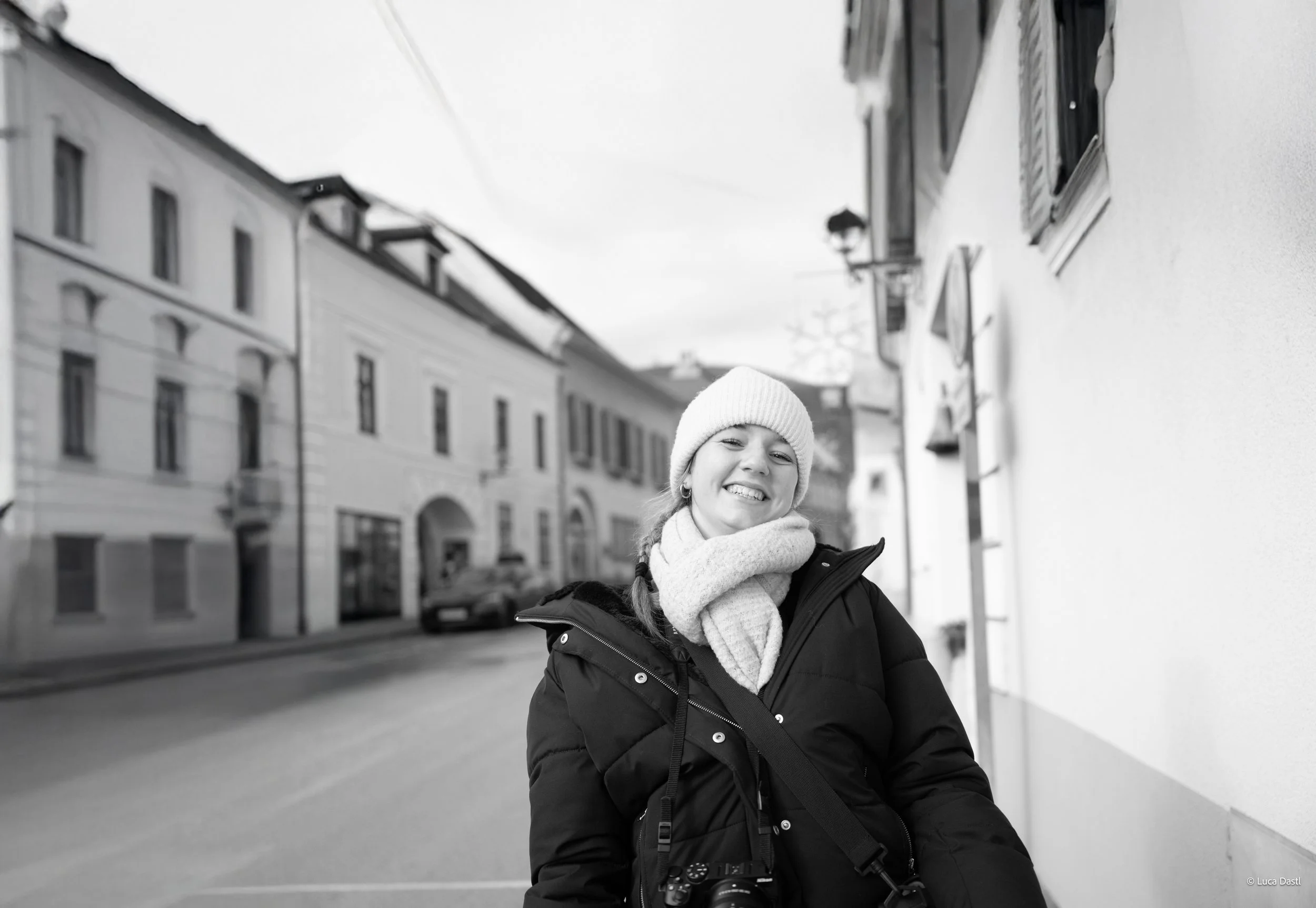 Junge Frau in Winterkleidung mit Mütze, Schal und Kamera lächelnd auf Stadtstraße in Deutschland, schwarz-weiß.