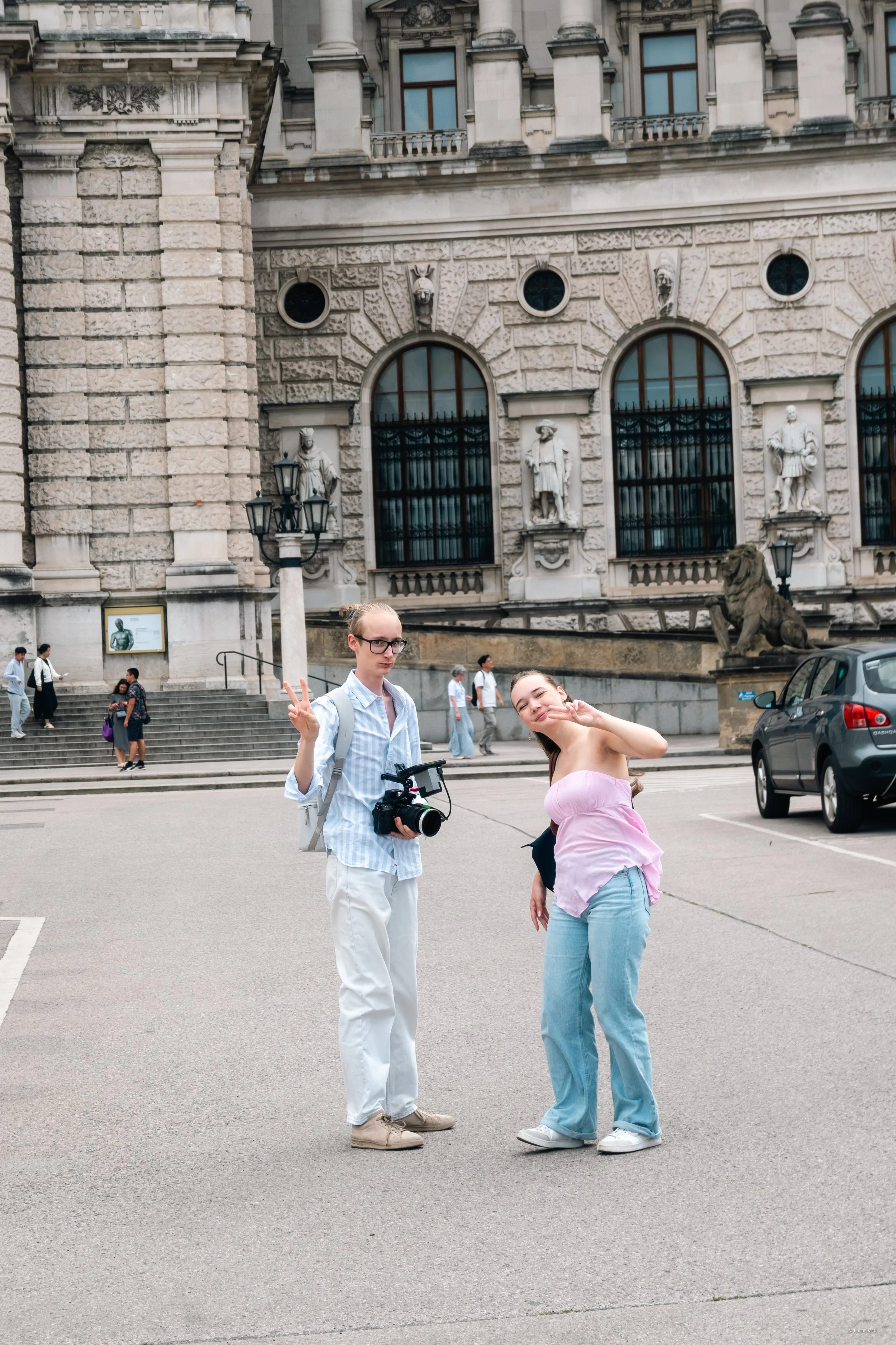 Zwei junge Frauen posieren vor einem historischen Gebäude, das typische Steingebäudefassaden und Skulpturen an der Fassade hat. Die eine Frau trägt eine rosa Top und blaue Jeans, die andere hält eine Kamera und macht eine Handgeste.