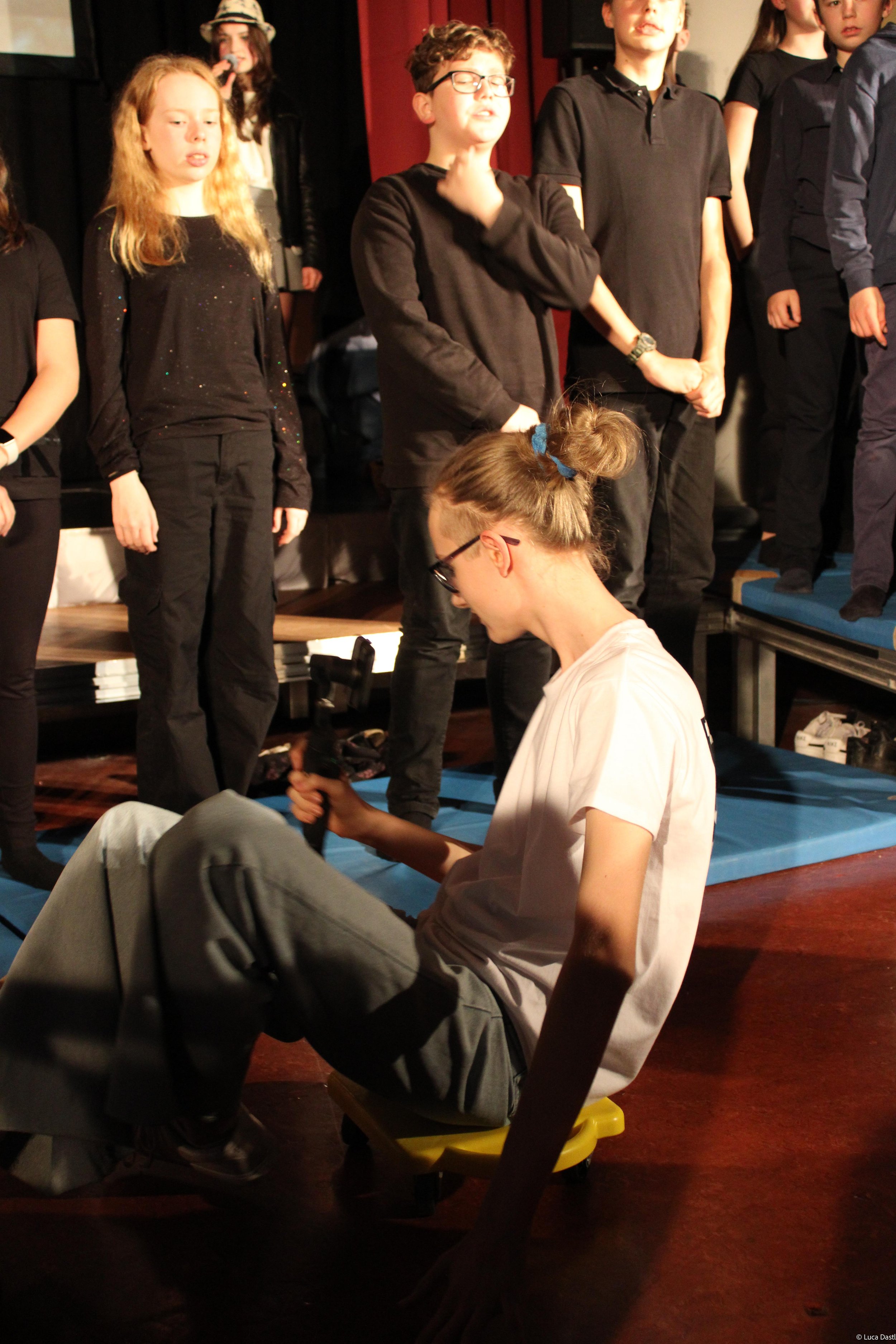 Jugendliche auf einer Bühne, einige stehen in einer Reihe, eine sitzt auf einem Rollstuhl im Vordergrund, alle scheinen Teil eines Theaterstücks oder einer Revue zu sein.