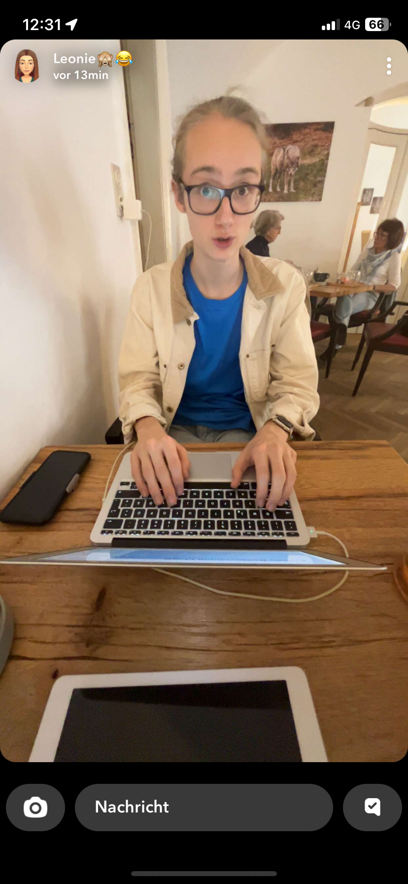 Junger Mann mit Brille und blonder Haare arbeitet an einem Laptop in einem Café, im Hintergrund sitzen zwei Frauen und es hängt ein Gemälde eines Pferdes an der Wand.