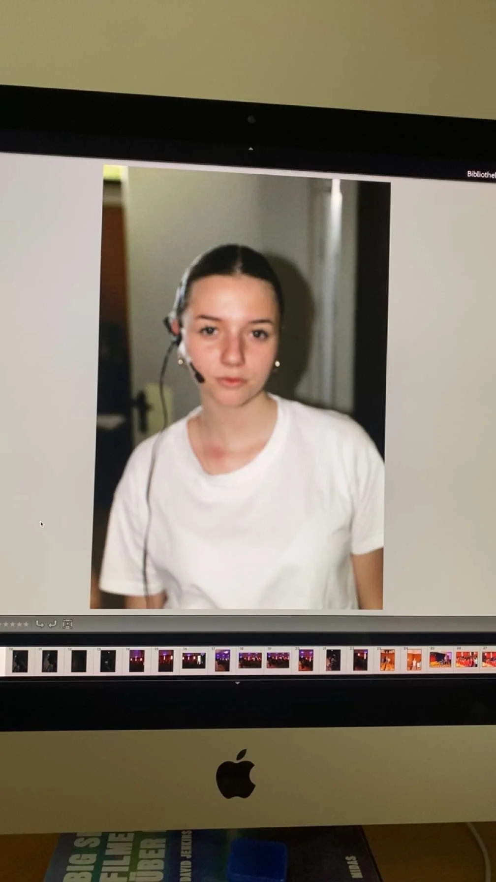 Computerbildschirm zeigt ein Porträt einer jungen Frau mit dunklen Haaren in einem weißen T-Shirt, trägt ein Headset, im Hintergrund eine weiße Wand mit Tür.