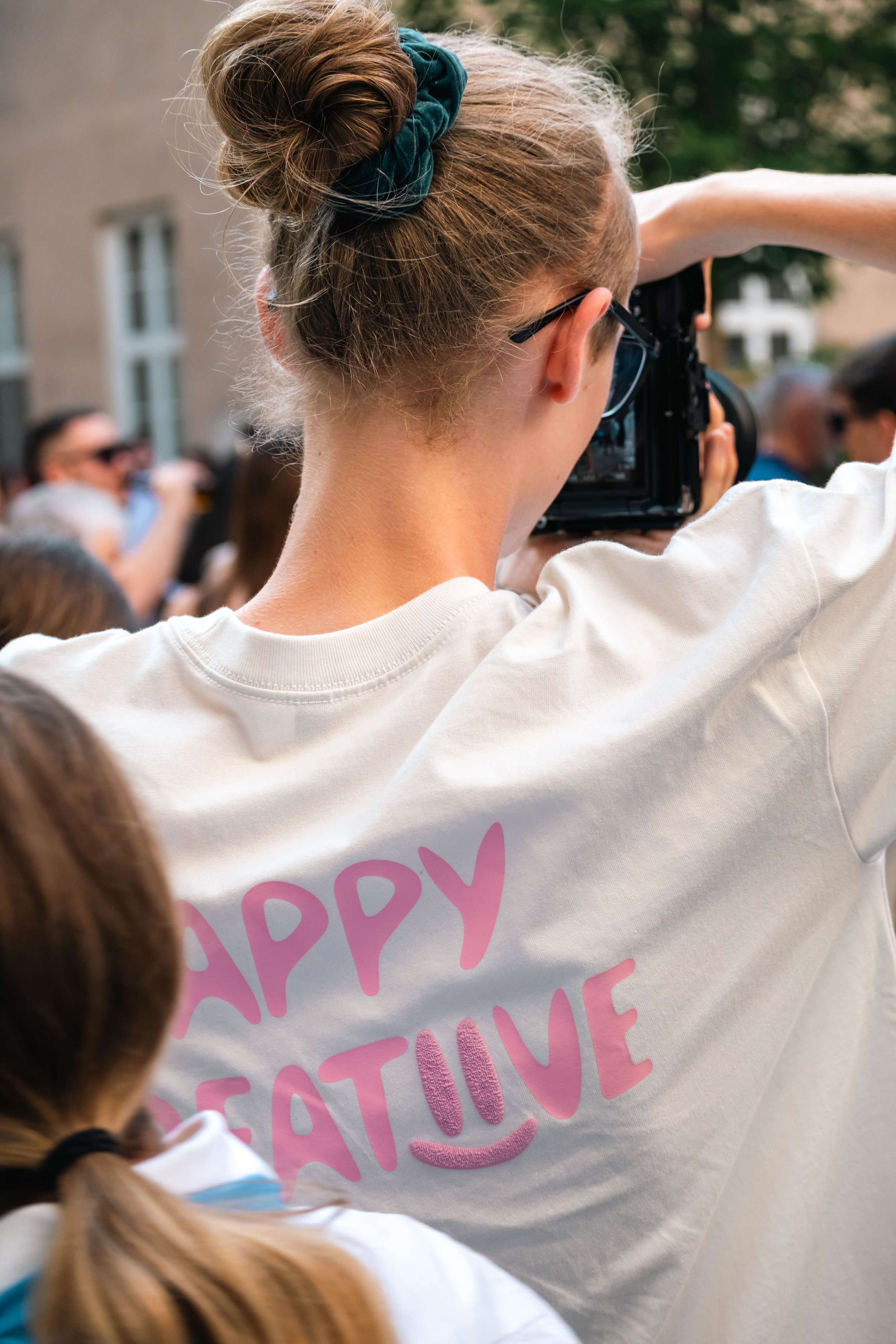 Frau mit braunen Haaren in Zopf, trägt ein weißes T-Shirt mit pinker Aufschrift 'HAPPY GATEINE' und macht Fotos bei einem öffentlichen Event.