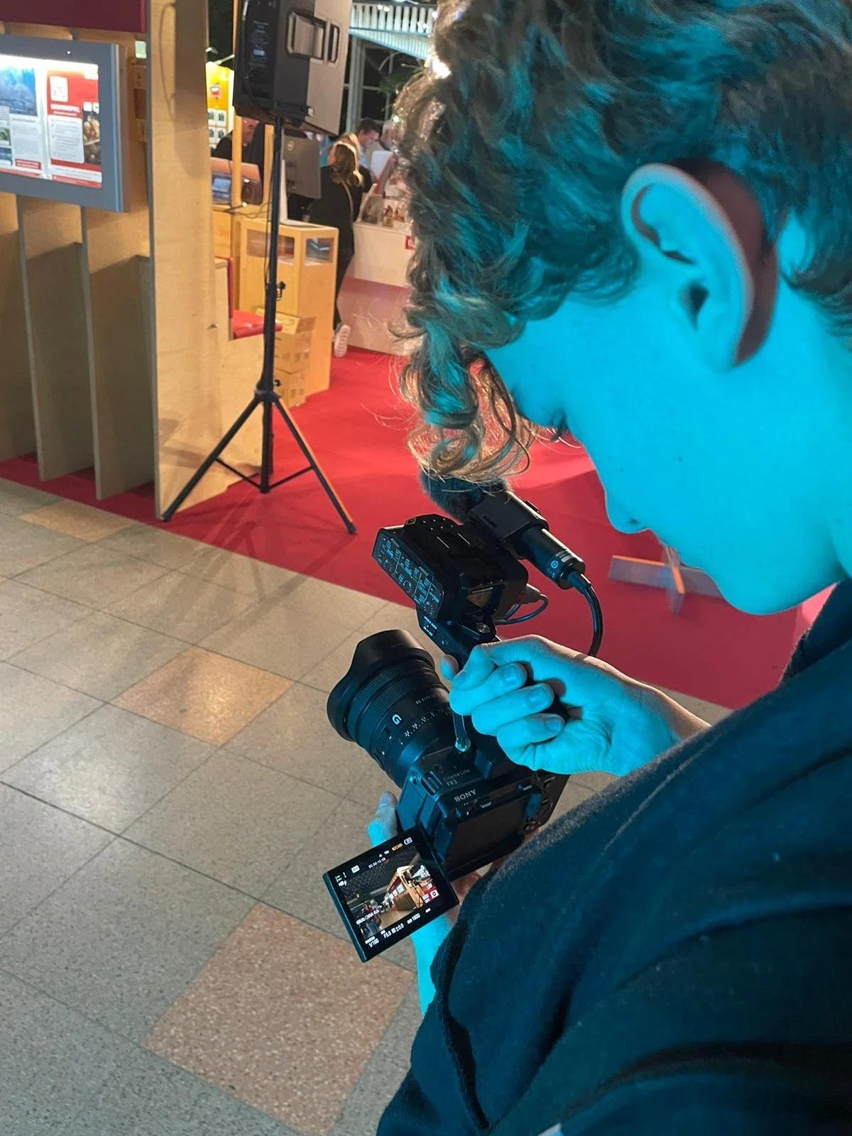 Ein junger Mann mit Lockenfil hair, der eine Kamera hält und fotografiert. Im Hintergrund sind Menschen und eine Verkaufsstand mit roten Teppichen.