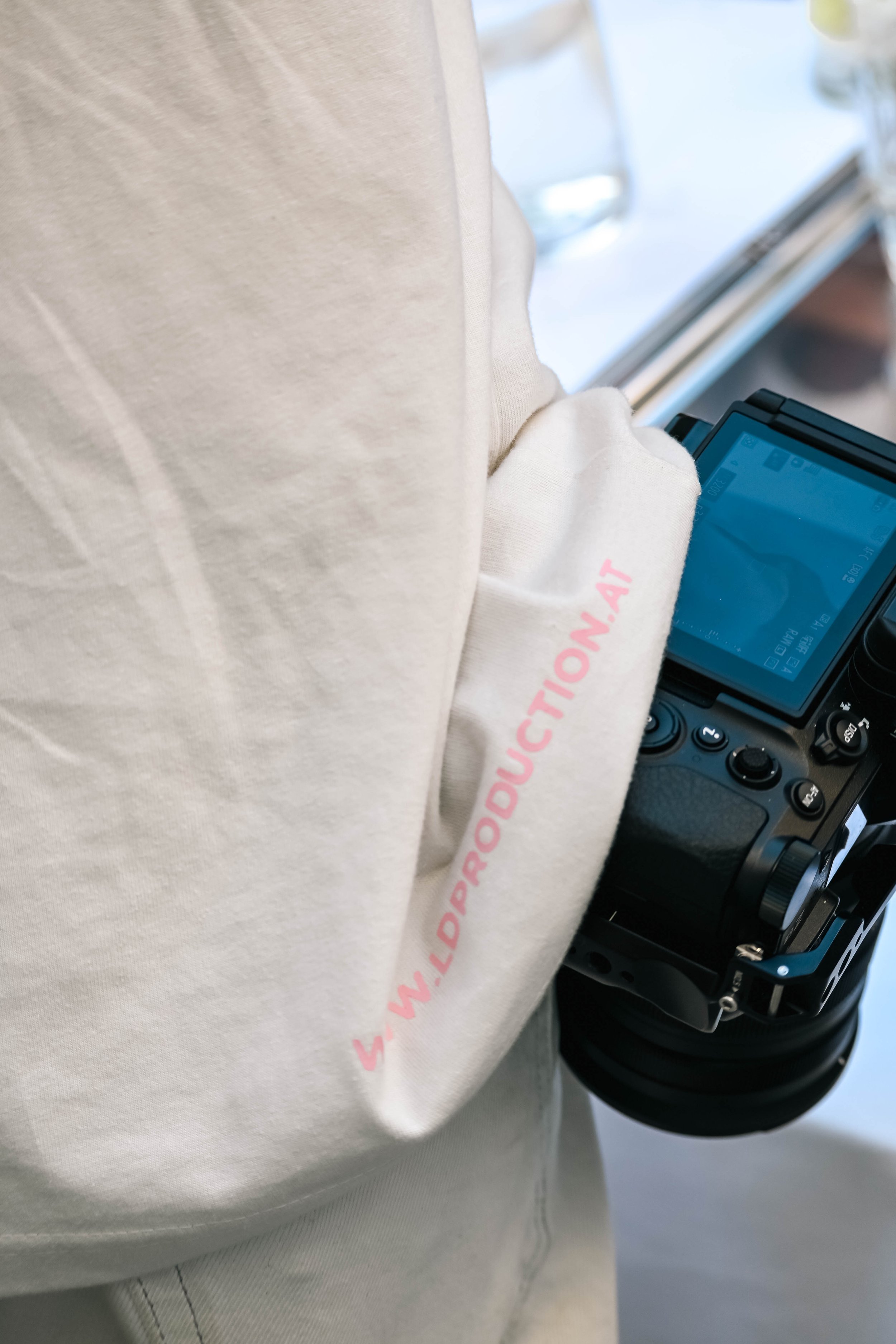 Person trägt weiße Sweatjacke mit pinkem Aufdruck auf dem Ärmel, hält eine professionelle Kamera.