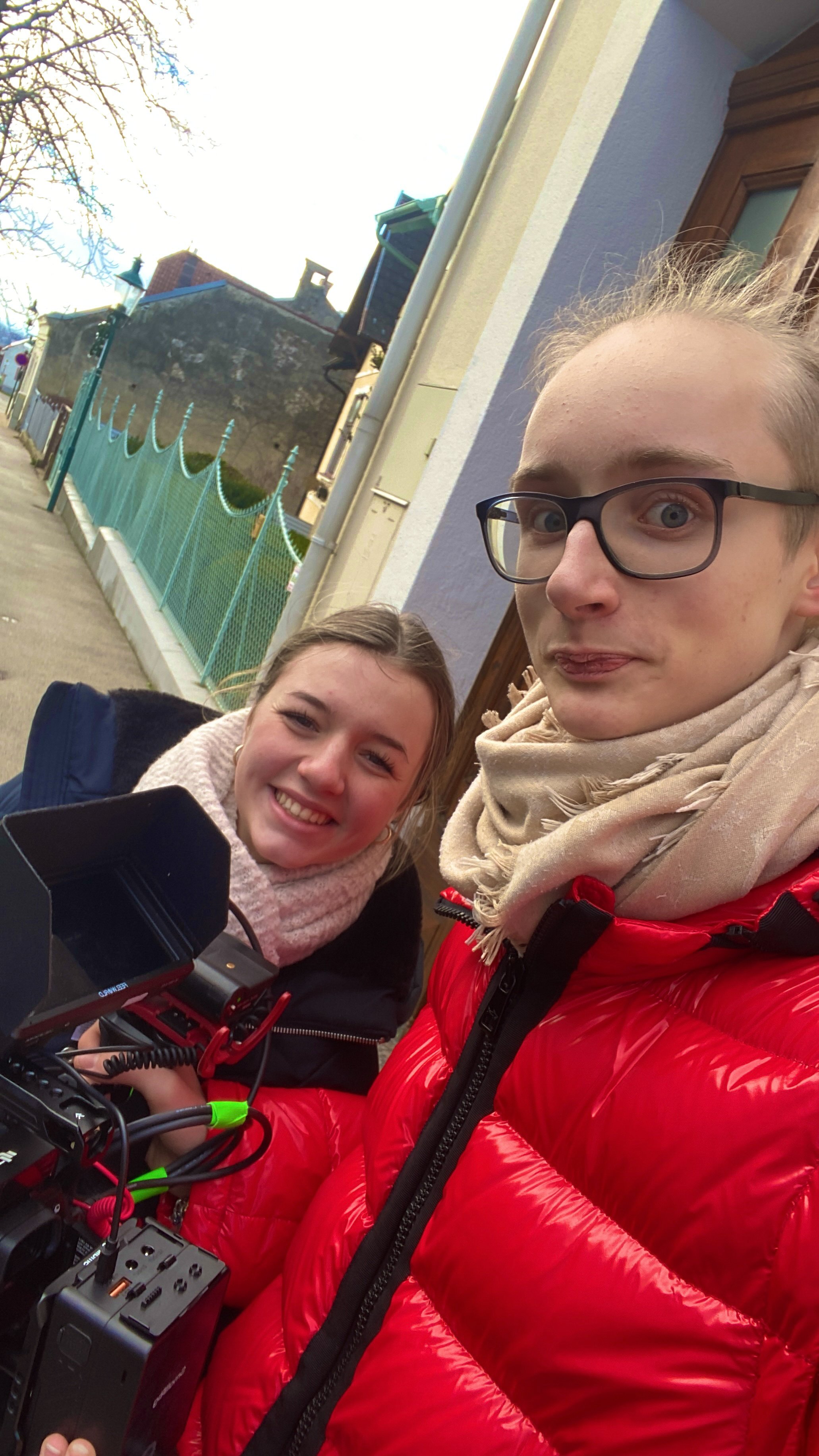 Zwei junge Menschen, eine Frau und ein Mann, posieren für ein Selfie auf einer Straße. Die Frau lächelt, der Mann trägt Brille und eine beige Schal, trägt eine rote Jacke. Es ist ein sonniger Tag, im Hintergrund sind Häuser und eine Bahnseite sichtba