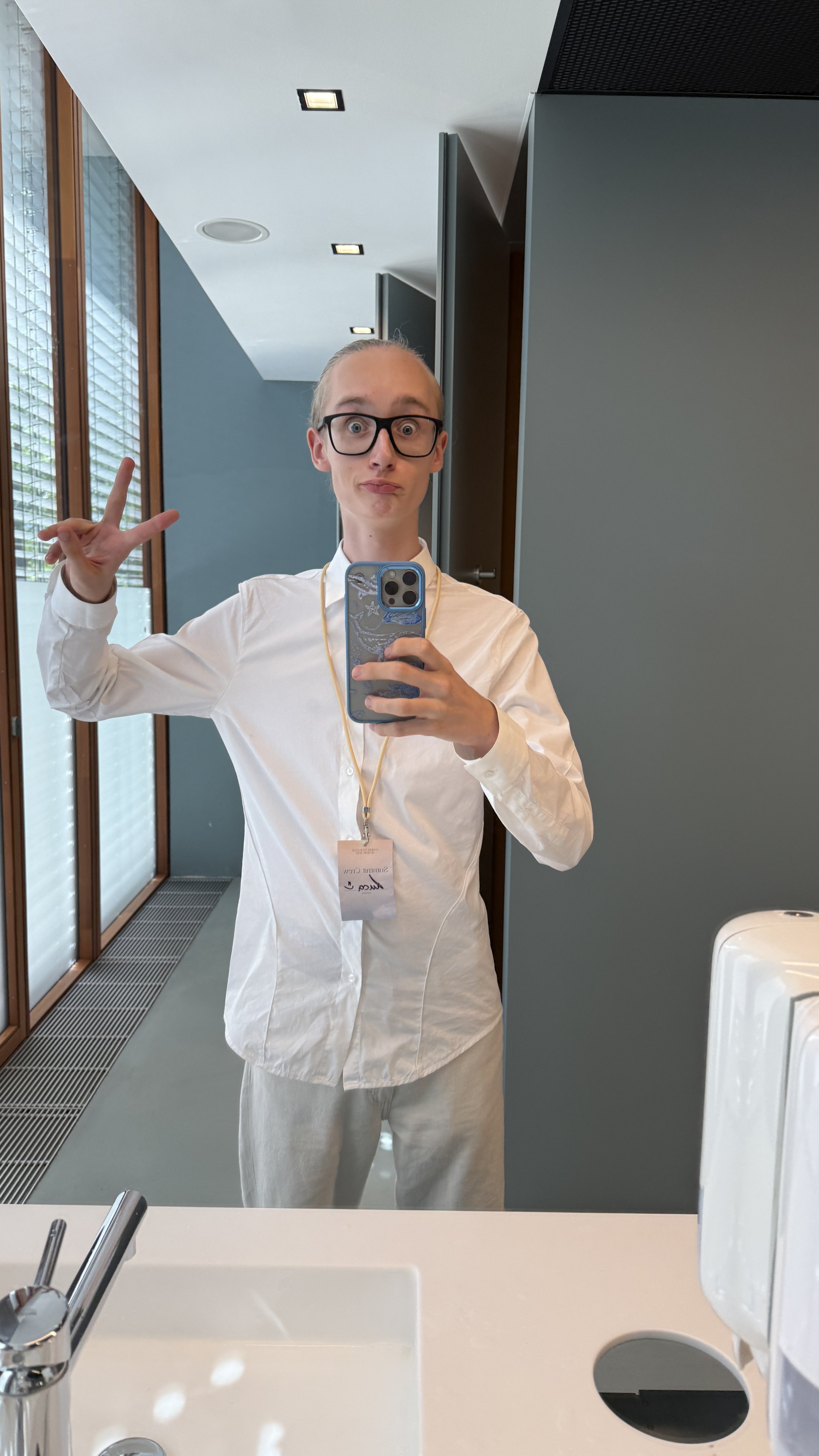 Junger Mann mit Brille und weißen Hemd, macht Peace-Zeichen und nimmt Selfie im Badezimmer, hinter ihm Fenster mit Jalousien, im Hintergrund graue Wände