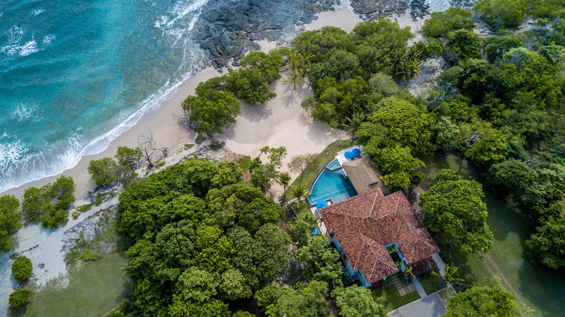 Aerial view of a tropical beach house surrounded by lush trees, with a swimming pool, sandy beach, and ocean waves nearby.