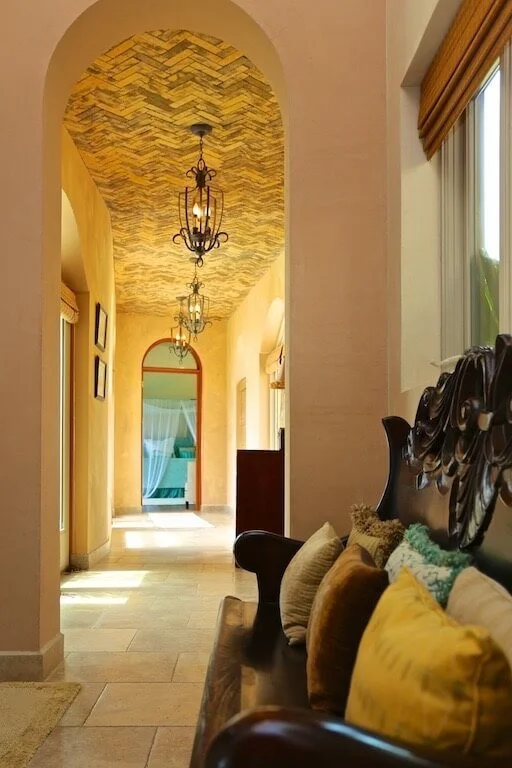 A hallway in a house with a barrel ceiling featuring a decorative herringbone pattern and wrought iron chandeliers. Light from a window on the right illuminates the space. A wooden bench with colorful pillows is visible on the right side.