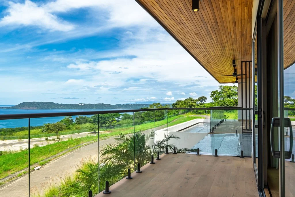 Modern balcony with glass railing overlooking green landscape, water, and sky, part of a contemporary building with wood-paneled ceiling.