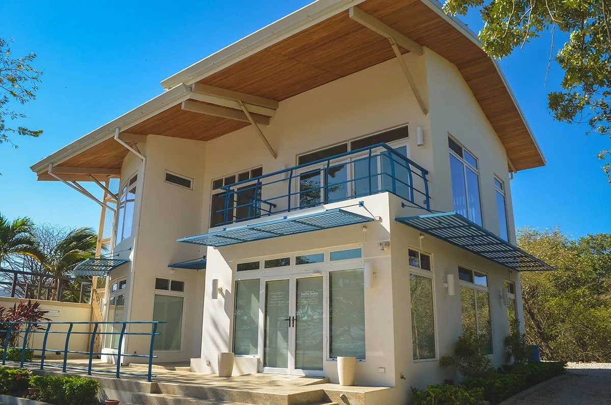 Modern two-story house with large windows, glass balcony railings, a curved roof, and surrounding greenery under a clear blue sky.