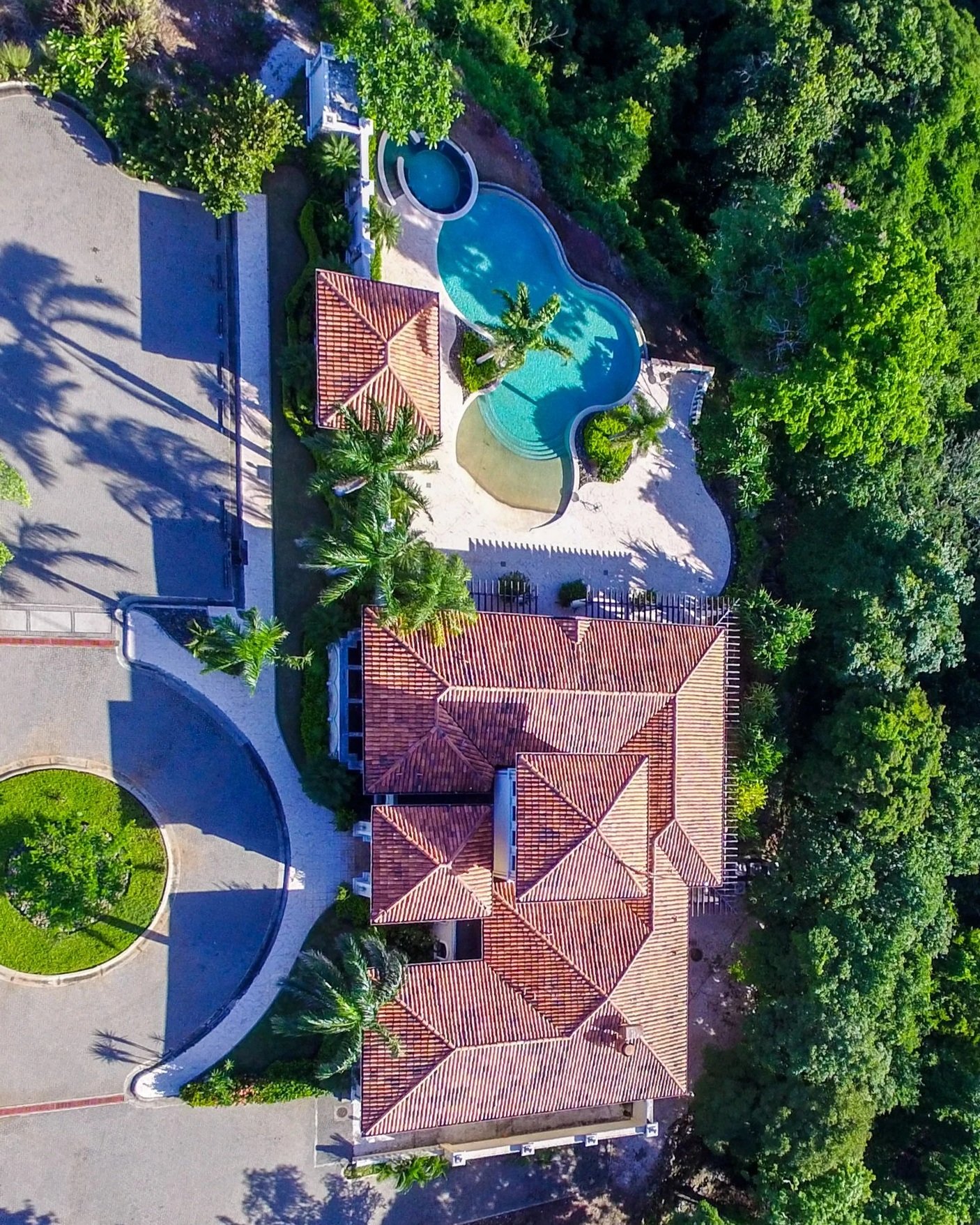 Aerial view of a house with a red tile roof, swimming pool, and lush greenery surrounded by trees.