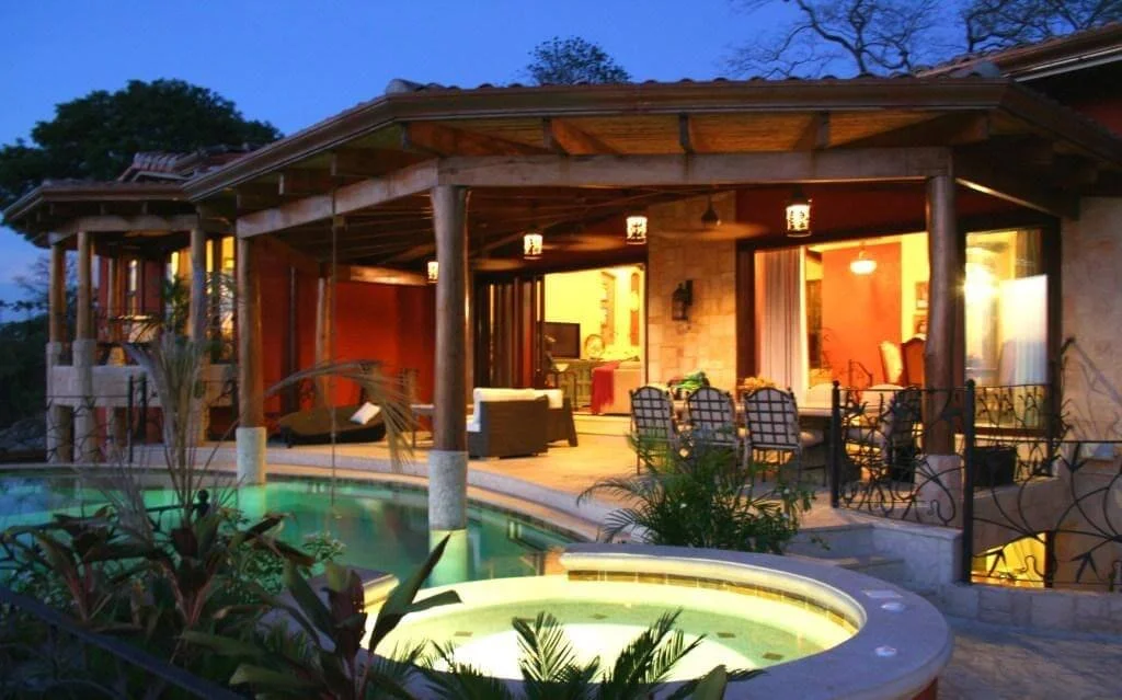 A luxurious outdoor patio with a swimming pool, lounge chairs, and a covered seating area illuminated by warm lighting, attached to a house at dusk.