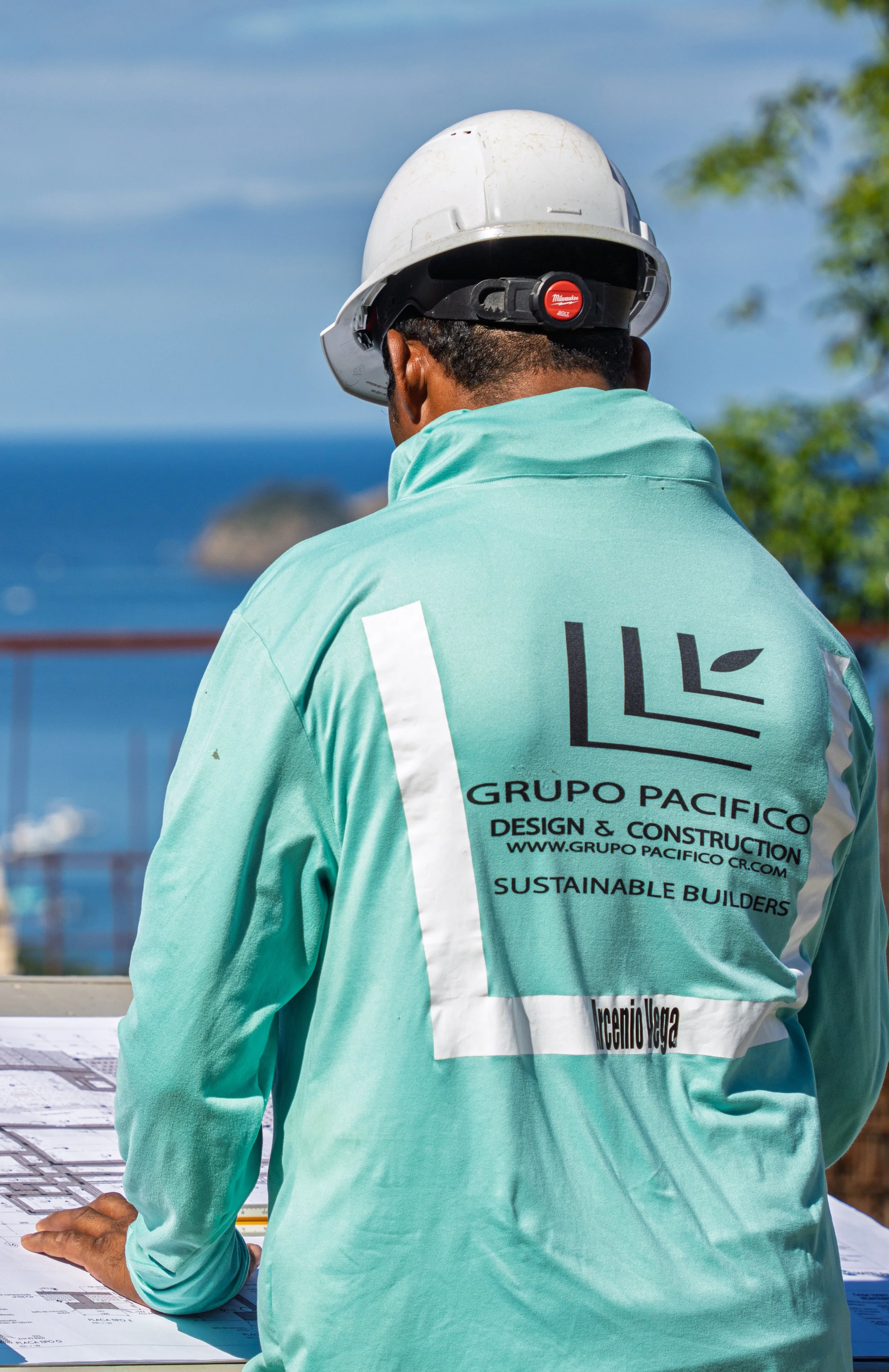 A construction worker wearing a white safety helmet and a light blue shirt with the logo and name of Grupo Pacifico Design & Construction, standing outdoors, looking at blueprints on a table, with a background of blue sky, ocean, and some green trees.