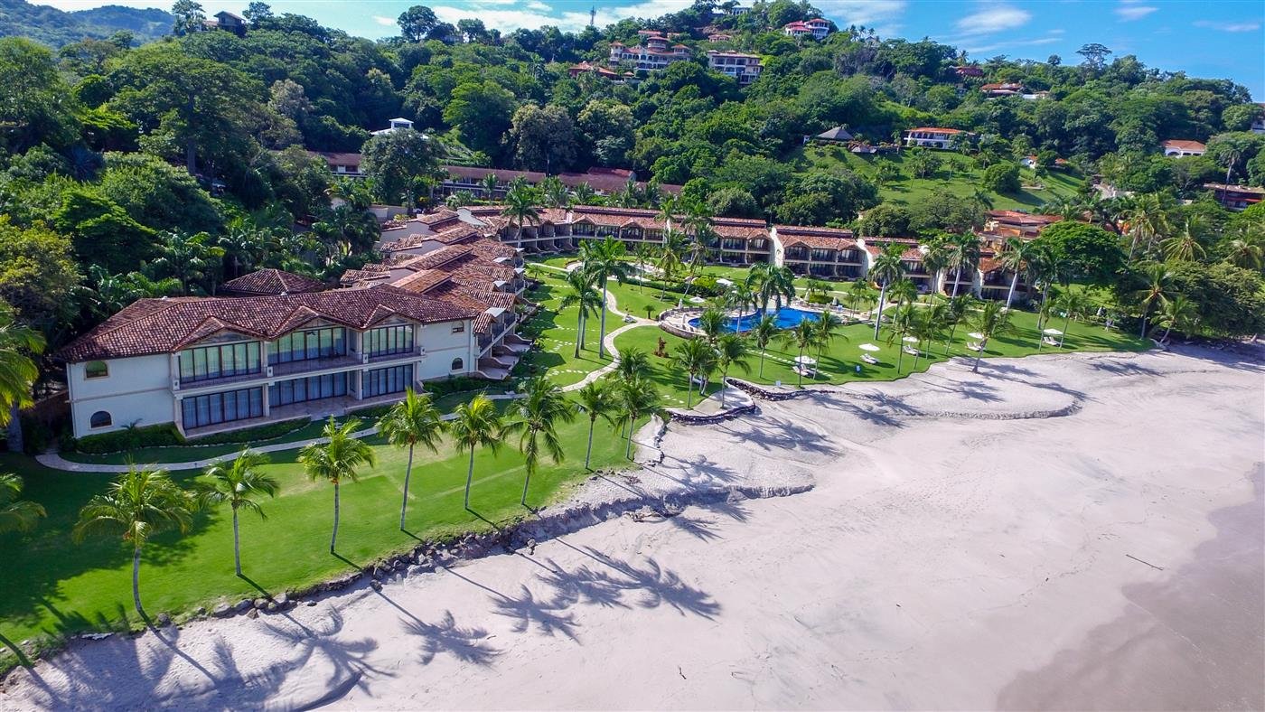 Aerial view of a beachside resort with white buildings featuring red-tiled roofs, a swimming pool, and green landscaped lawns with palm trees, overlooking a sandy beach and the ocean.