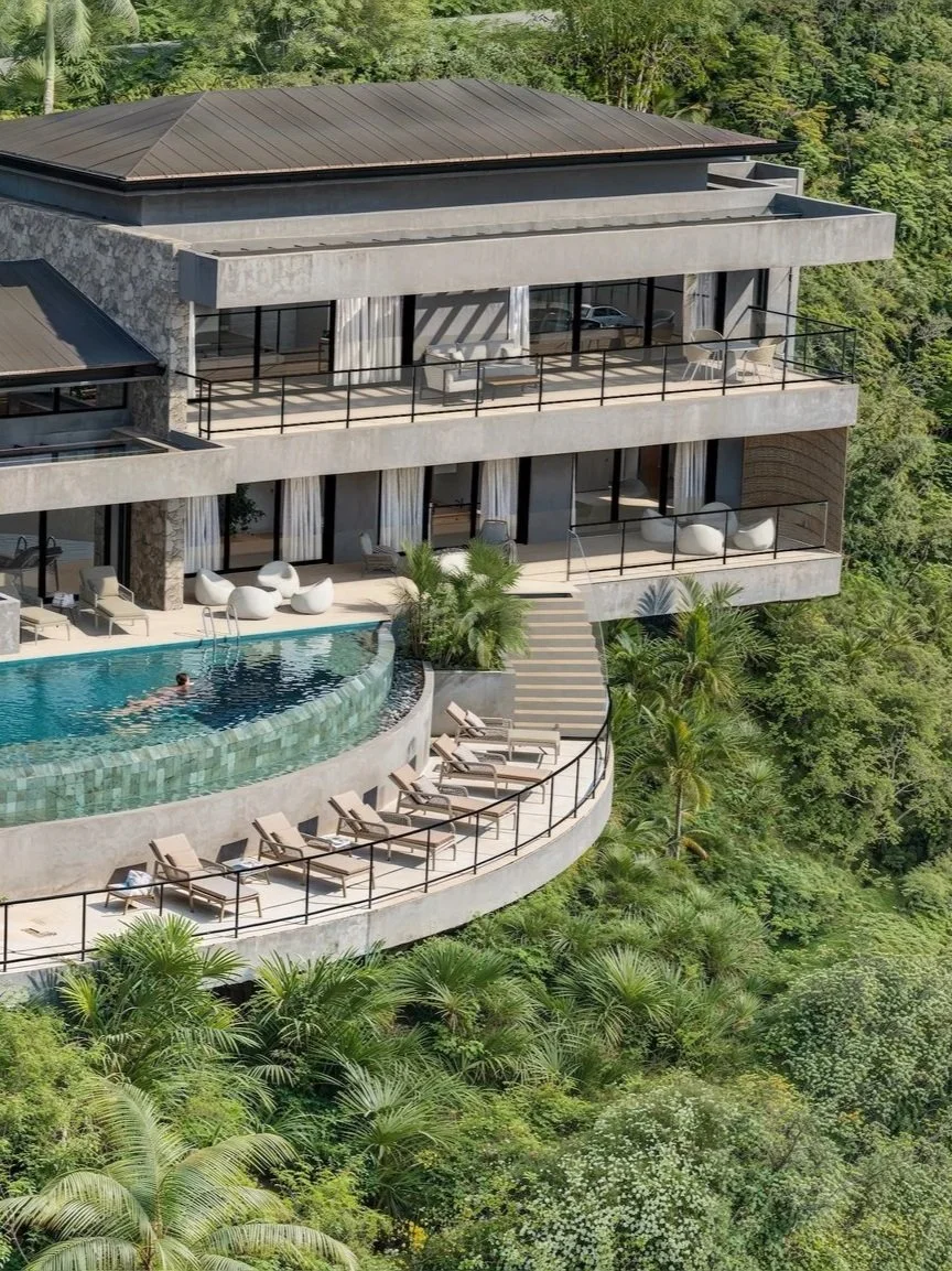 Modern multi-story house with a swimming pool on a balcony, surrounded by lush green trees.